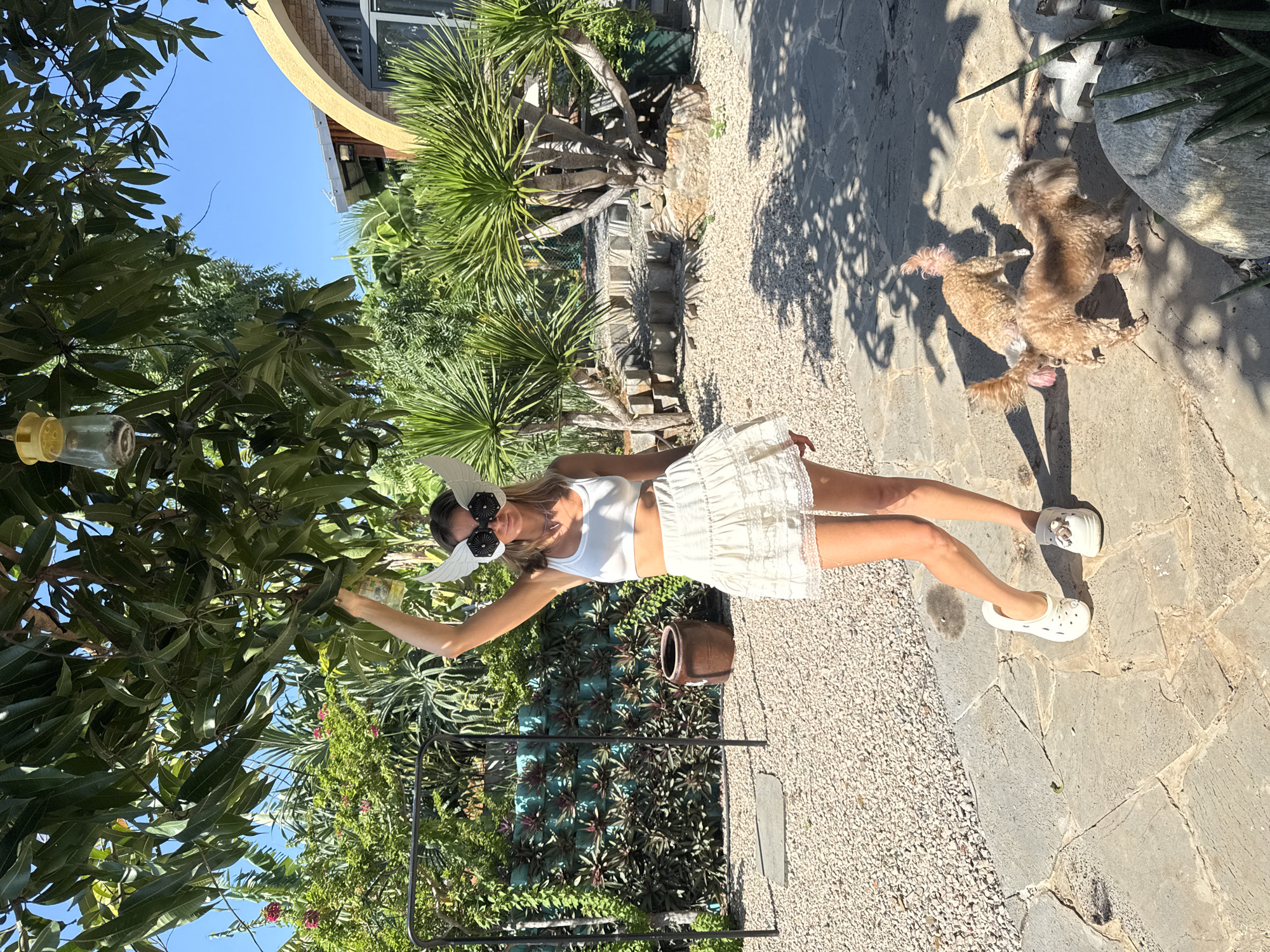 A woman with blond hair, wearing a white crop top, a short white ruffled skirt, and white Crocs, poses outdoors in Mũi Né, Vietnam. She has on large, white winged eye mask with black accents and sunglasses. She is holding a bottle in her right hand above her head. Two small, tan dogs are in the foreground, near her feet. The woman stands on a stone patio, surrounded by gravel and lush greenery, including palm trees and a large tree with a bird feeder hanging from its branches. In the background, a building with a yellow archway is visible. The scene is brightly lit by sunlight, casting shadows on the ground.