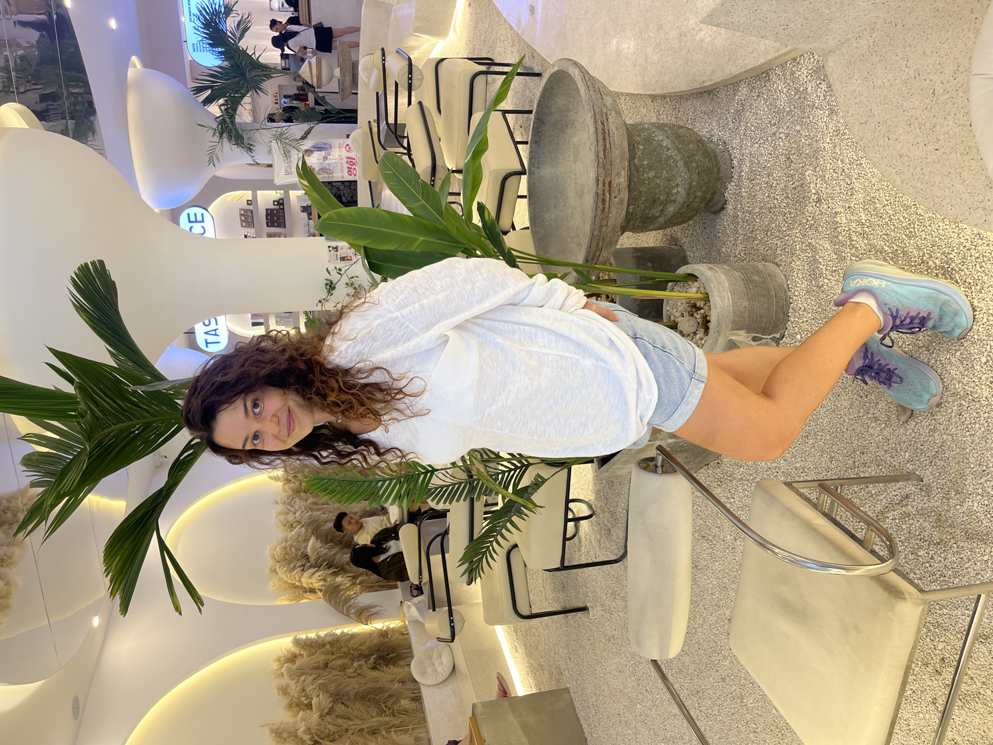 A woman with curly brown hair, wearing a white long-sleeved shirt and denim shorts, stands in a cafe in Mũi Né, Vietnam. She is facing the camera with a slight smile. Behind her, large potted plants and decorative dried grass add to the tropical ambiance. The cafe features a modern, minimalist design with white curved walls and surfaces, and light-colored flooring. Several chairs, some upholstered in cream and with chrome frames, are arranged around the space. In the background, a counter with products and other patrons can be seen.