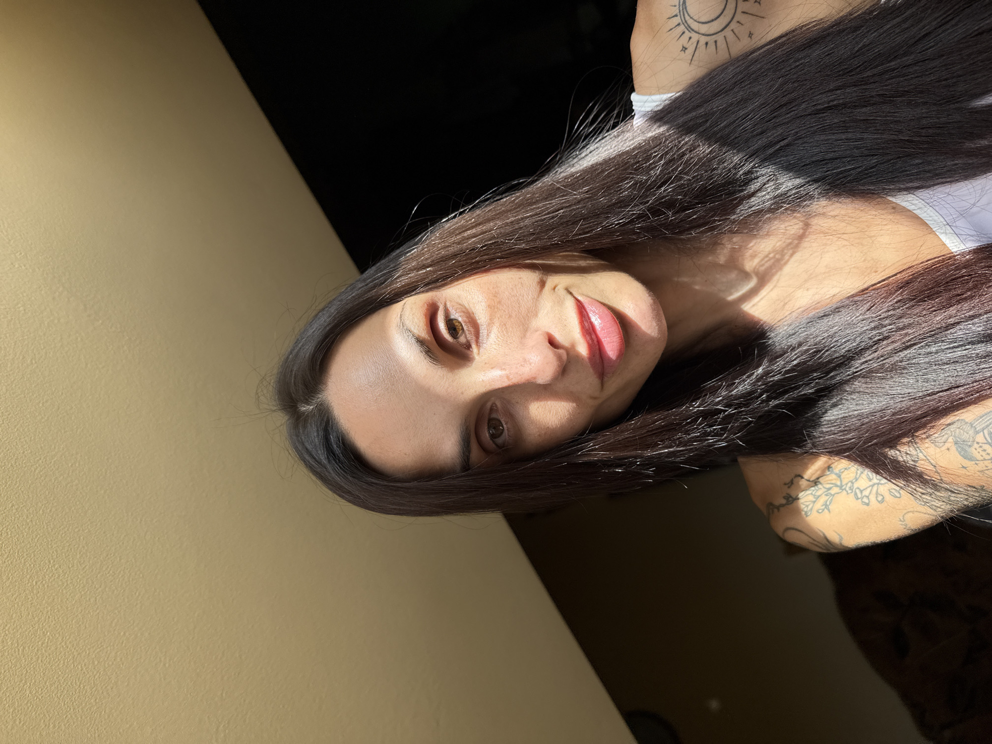 This media file presents a close-up portrait of a woman. She has long, dark, straight hair, which is illuminated by strong, direct sunlight. Her face is turned slightly towards the viewer, featuring light brown eyes and a relaxed, subtle smile with her lips gently parted. The bright sunlight creates prominent highlights on her forehead, cheekbones, nose, and lips, as well as catching individual strands of her hair, making them shine. In contrast, other areas of her face and the background are cast in deep shadow, contributing to a high-contrast aesthetic.
The woman is wearing a light-colored, possibly white, top. Several tattoos are visible on her right side; one on her upper chest/shoulder depicts a celestial motif with a stylized sun, moon, and stars, while another on her arm features delicate floral or organic patterns. The background suggests an indoor environment, with a plain, light yellowish-beige wall on the left side of the frame and a dark, indistinct area on the right.
The intense, directional light strongly indicates a daytime setting. The woman's expression appears calm and gently engaging. The provided context states this image is from Pleven, Bulgaria.