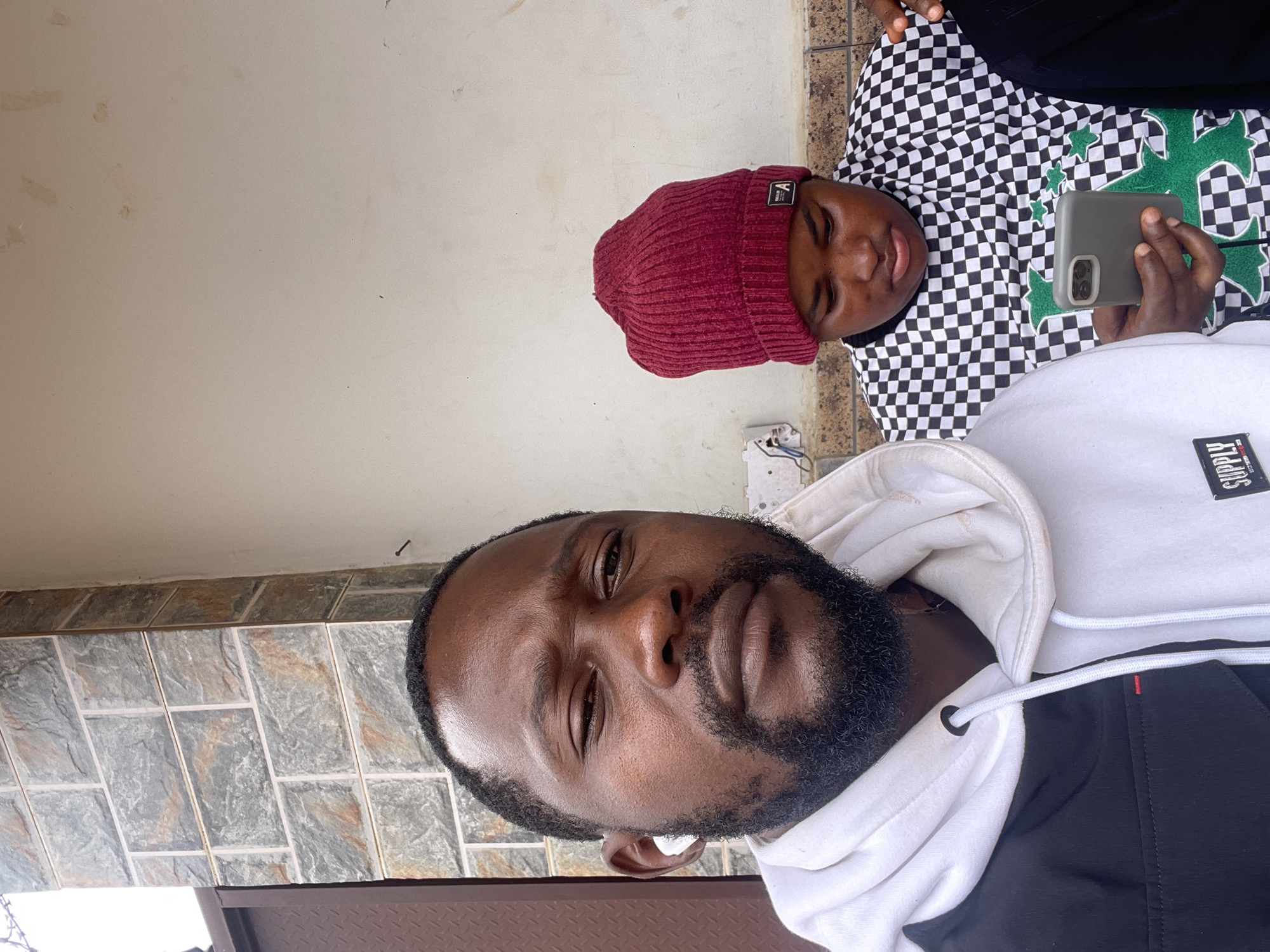In Kurunduma, Nigeria, a selfie is taken outdoors, showing two individuals. The person in the foreground, a man with dark skin, a beard, and a white earbud in his left ear, faces the camera. He is wearing a white hoodie with black accents. To his right, another person, wearing a red knitted beanie and a black and white checkered shirt with a green graphic, looks down at a smartphone held in their hand. The background features a textured brick wall and a plain wall. The overall scene suggests a casual, possibly urban, setting in Nigeria.