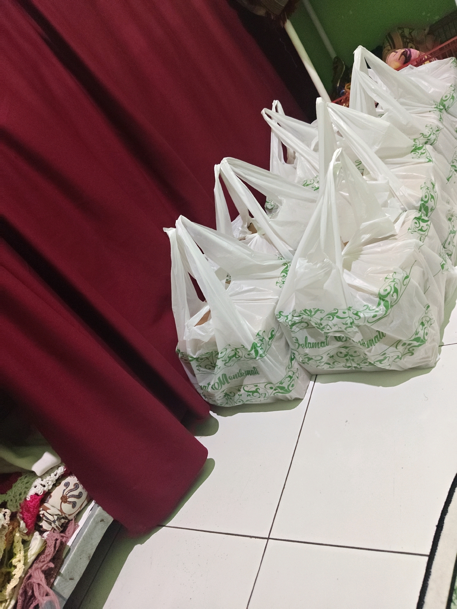 In Sumberwono, Indonesia, a collection of white plastic bags with green decorative trim and Indonesian text, likely "Selamat Menikmati" (Enjoy Your Meal), are arranged in a staggered fashion on a tiled floor. The bags are filled, suggesting they contain food items or take-away orders. A deep red curtain occupies the left side of the frame, creating a backdrop for the bags. To the right of the bags, a sliver of a green surface is visible, possibly a shelf or wall, with what appears to be fabric or blankets stacked on it. In the lower left corner, a glimpse of colorful woven material is visible, peeking out from under the curtain. The lighting suggests an indoor setting, possibly a home or small business.