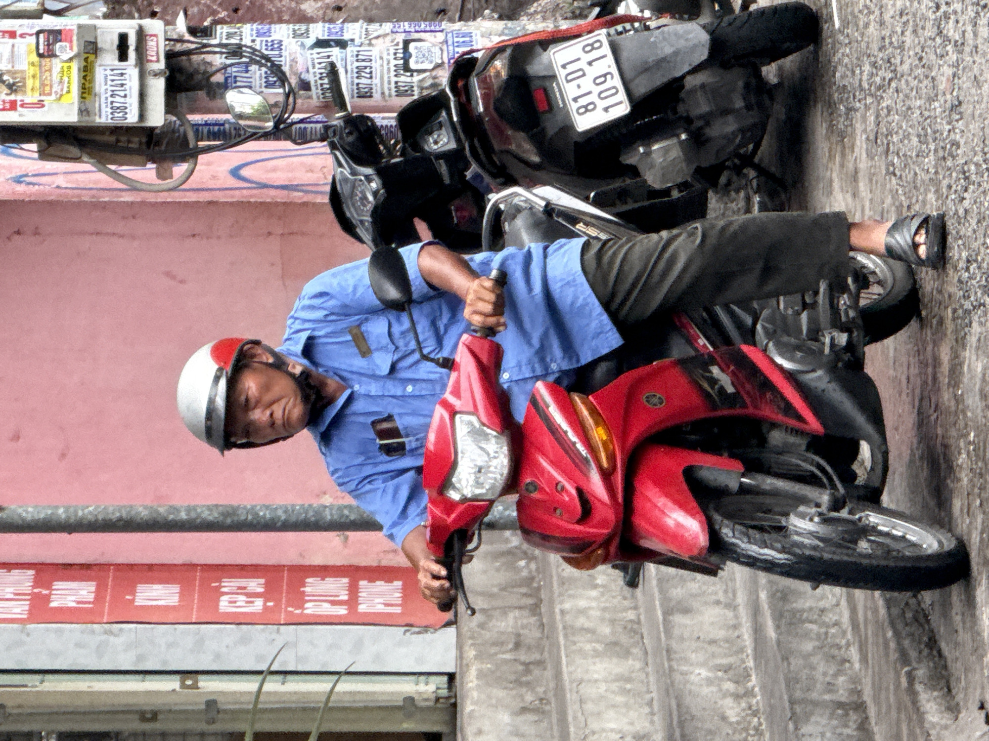 The image captures a street scene in Nha Trang, Vietnam, focusing on a man riding a red and black scooter. The man, wearing a white and red helmet and a blue shirt, is seated on the scooter, with his left hand on the handlebars. His right hand is not visible. He is wearing dark pants and dark sandals. The scooter is red in the front and black in the rear, with visible handlebars, mirrors, and a license plate that reads "81-D1 109.18".
Behind the man and his scooter, a pink wall serves as a backdrop. To the left of the man, a vertical red sign with white text is partially visible. To the right, a utility pole with various notices and stickers is present. The ground appears to be wet, suggesting recent rain. Steps made of concrete are visible on the left side of the image. The overall impression is a candid snapshot of daily life on a street in Vietnam.