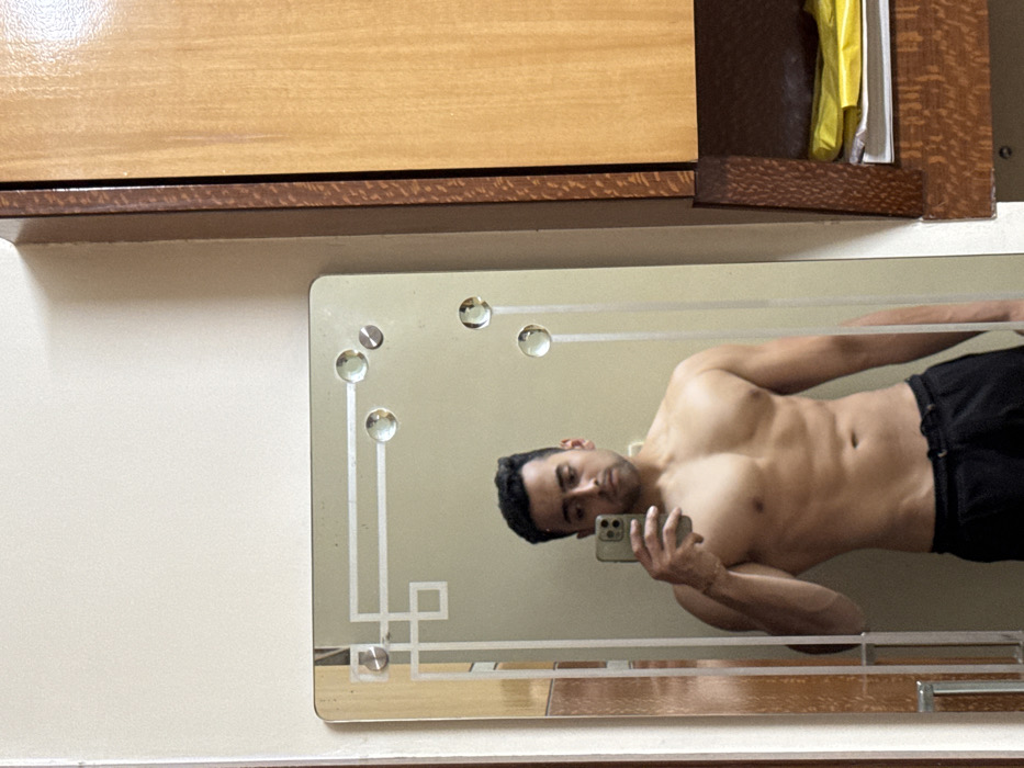 A shirtless man takes a mirror selfie. He holds a phone in his left hand and angles his body toward the mirror. His face is visible. He wears dark-colored pants or shorts. The mirror is rectangular with decorative edges and circular accents. It hangs on a light-colored wall beneath wooden cabinets. Some items are partially visible in the cabinet above the mirror.