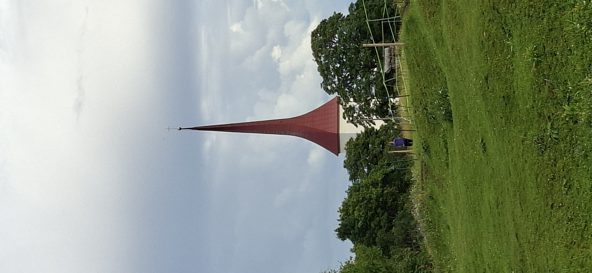 The image depicts a rural landscape with a prominent church spire in the background. The spire is tall and conical, painted a vibrant red, and topped with a cross. It rises from a white church building, partially obscured by lush green trees. In the middle ground, a person wearing a purple top and dark pants stands on a grassy slope, facing away from the camera. A simple wooden fence with green netting runs horizontally across the scene, separating the grassy area from the higher ground where the church is situated. The foreground is dominated by a rolling green meadow, dotted with wildflowers and patches of darker vegetation. The sky is overcast with dramatic clouds, suggesting either impending rain or the aftermath of a storm. The overall impression is one of natural beauty and tranquility. This church is likely the Rakvere Church of the Holy Trinity, a historic Lutheran church located in Rakvere, Estonia.