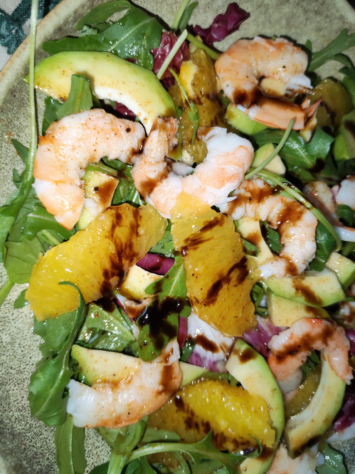 A close-up shot captures a vibrant salad served in an earth-toned, speckled ceramic bowl. The salad consists of several key ingredients: cooked, shelled shrimp, slices of ripe avocado, bright segments of orange, and a generous bed of mixed greens, including dark green arugula and some reddish-purple lettuce leaves.
The shrimp are a light pinkish-orange, visibly seasoned with small dark specks, suggesting pepper or other spices. The avocado slices are pale green with a slightly yellowish hue towards the center, appearing creamy and fresh. The orange segments are peeled clean of pith, revealing their juicy, yellow-orange flesh. These components are scattered over the fresh, crisp greens.
A dark, glossy, and viscous dressing, possibly a balsamic glaze or reduction, is artfully drizzled over the entire salad, creating dark streaks and pools that contrast with the lighter colors of the ingredients. The presentation suggests a carefully prepared meal. The lighting appears to be indoor, highlighting the textures and colors of the food. No people or visible text are present in the image.