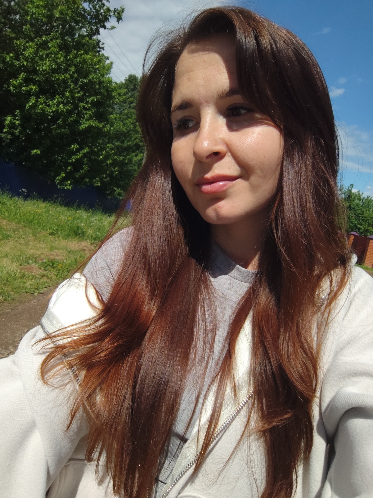 A woman is outdoors in Фоки, Russia. She has long, reddish-brown hair and is wearing a gray shirt under a light-colored zip-up jacket. Behind her is green foliage, a blue fence, and a blue sky with light clouds.