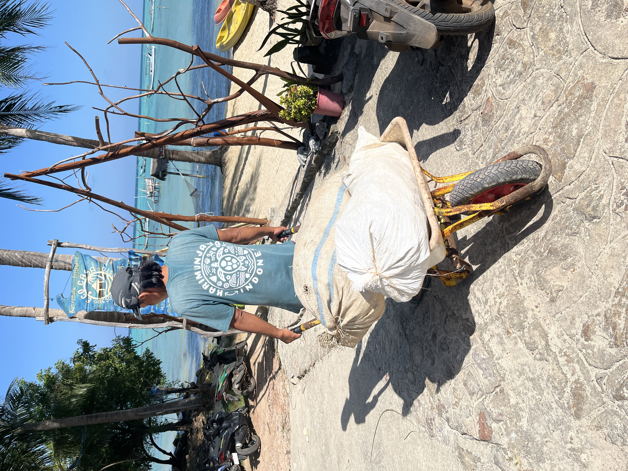 A person wearing a dark cap and a teal t-shirt with a white circular graphic and text walks away from the camera, pushing a yellow wheelbarrow filled with two large sacks. The sacks are made of rough, light-colored material and are bound with rope. The person is on a textured, grey stone pathway. To the left of the pathway, there are several parked motorcycles and lush green trees. Behind the person and the wheelbarrow, a bright blue ocean meets a sandy beach, dotted with palm trees and some wooden structures. In the distance, several boats are visible on the water. The sky is clear and blue, indicating a sunny day. The scene appears to be a tropical coastal area, possibly in Legonipah, Indonesia, given the inscription on the person's shirt and the context of the location.