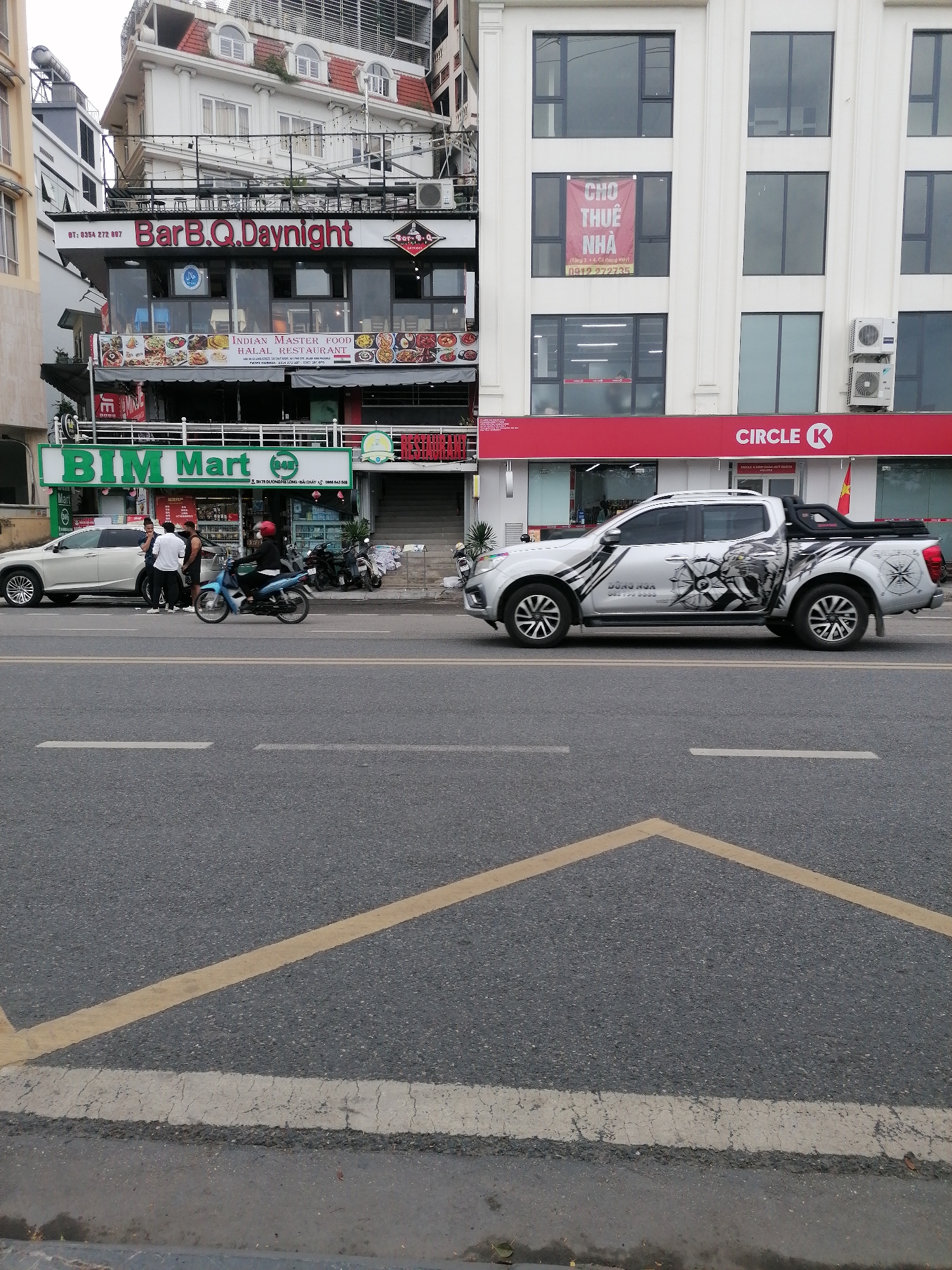 This image shows a street scene in Bai Chay Ward, Vietnam. In the foreground is an asphalt road with yellow lane markings. A silver pickup truck with distinctive graphics along the sides is parked next to a Circle K store. In the background, a building with a "BIM Mart" store on the ground level is visible, along with a "BarB.Q.Daynight" restaurant on the upper floors. Several people are gathered near the BIM Mart store, and a scooter is passing by. A sign in Vietnamese offering a house for rent is visible in the windows above the Circle K.