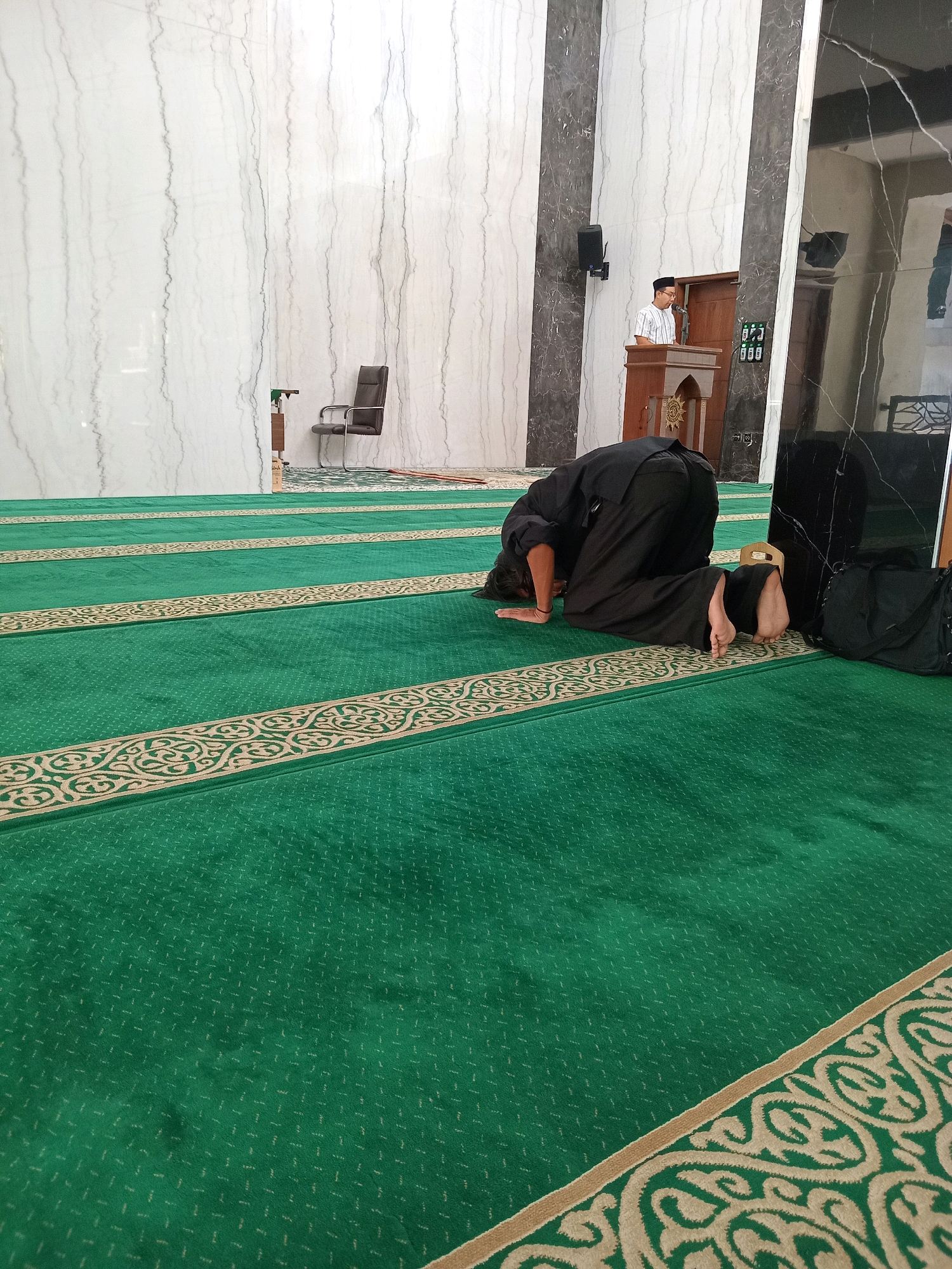 An exterior shot of a mosque in Randuagung, Indonesia captures a moment of prayer. The foreground is dominated by a vibrant green carpet with an intricate beige pattern, likely indicating rows for worshippers. In the midground, a man dressed in black is kneeling in prayer, his forehead touching the carpet, symbolizing submission. Further back, on a slightly elevated platform, another man stands near a wooden podium, presumably leading the prayer or delivering a sermon. The mosque's interior features white marble walls with subtle grey veins, contrasted by dark pillars. A black office chair is visible against one of the white walls, and a large black bag rests on the floor near the kneeling man. The overall scene depicts a serene and spiritual atmosphere within a modern mosque.