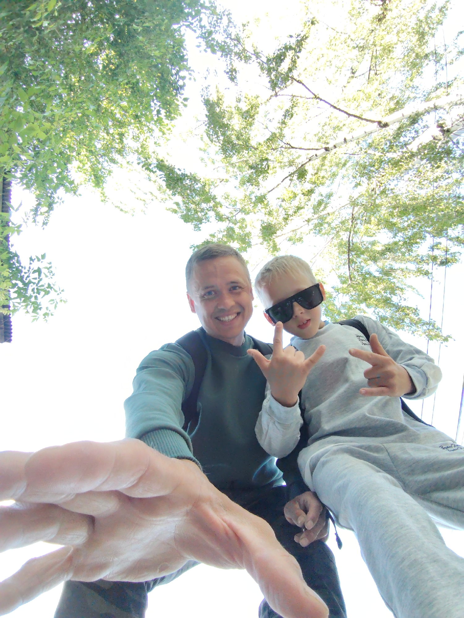 Captured from a low-angle perspective, the image features a man and a boy, likely father and son, posed beneath a canopy of green foliage. The bright sky illuminates the leaves, creating a somewhat overexposed backdrop.
The man, positioned to the left, wears a green long-sleeved shirt, a dark-colored backpack, and smiles towards the camera. His right hand is prominently displayed in the foreground, partially obscuring the view.
To the right, the young boy sports a gray tracksuit and dark sunglasses. He makes a "rock on" hand gesture with both hands. A black backpack is slung over his shoulders.
The location, Chaikovsky, Russia, is determined by the geolocation metadata attached to the image. However, the visible elements provide no further detail about the specific setting within the city. The primary focus is the interaction between the two subjects and their surroundings.
