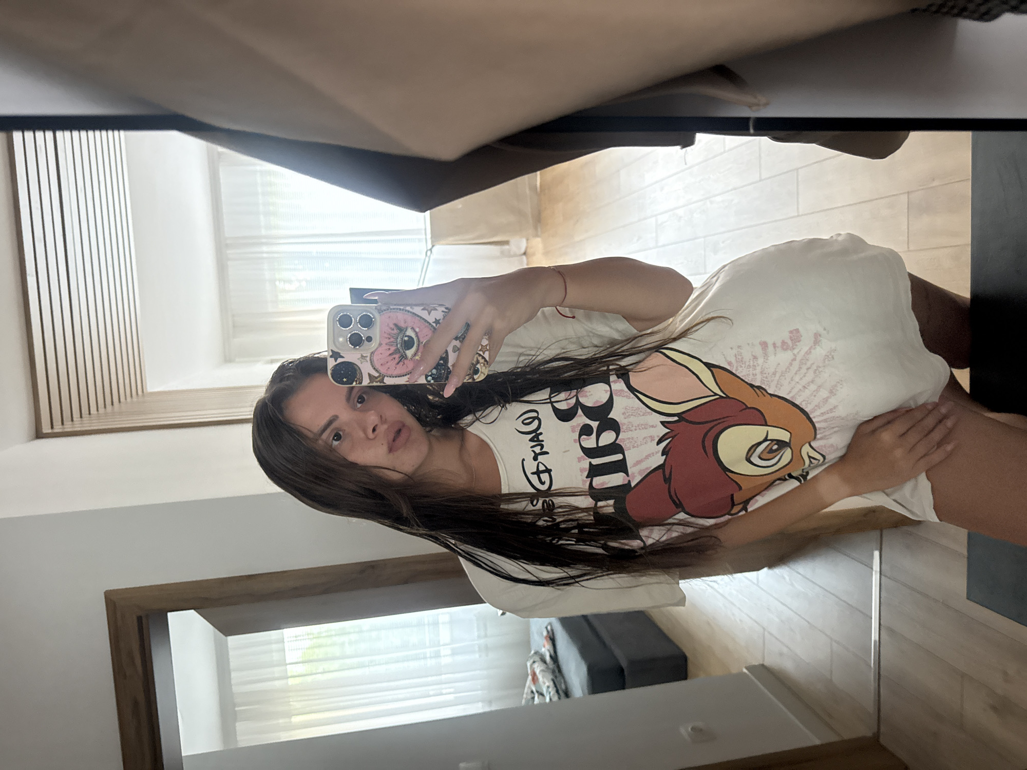 A young woman with long, dark, wet hair takes a mirror selfie in an indoor setting. She is wearing an oversized white t-shirt featuring a graphic of Bambi from Disney. Her right hand, holding a phone with a decorative case, is visible as she takes the photo. The background reveals a room with light-colored wooden floors, sheer white curtains, and a glimpse of a doorway leading to another area with a bed. The environment appears to be a residential interior, likely a bedroom or living space. There is no specific indication of this location being in Plovdiv, Bulgaria.