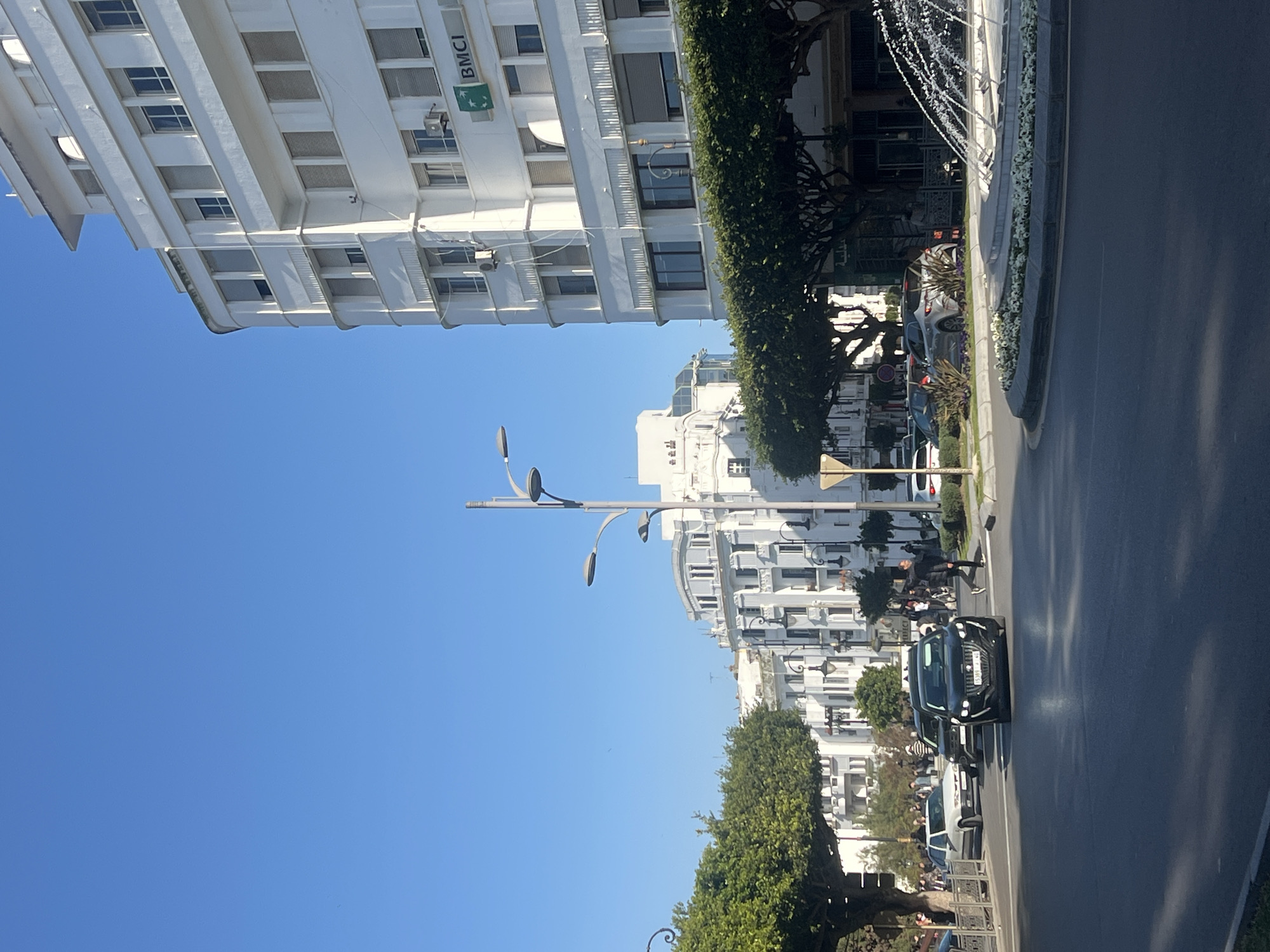 The image displays a sunlit urban street scene in Tanger (Tangier), Morocco, specifically near the intersection of Boulevard Pasteur and Rue de la Liberté.
On the right, a prominent multi-story white building, exhibiting Art Deco or Modernist architectural elements, dominates the frame. This building features multiple floors with rectangular windows, some fitted with external roller shutters, and displays the "BMCI" bank logo (green leaf design) on its lower facade. In the background, similar multi-story white buildings line the street.
The foreground and midground are occupied by a wide, paved street. A central circular island, acting as a roundabout, features a multi-tiered fountain actively spraying water. The island is landscaped with white flowers, primarily petunias, and green foliage. Several trees with dense green canopies line the street, casting shadows, particularly over an outdoor seating area on the right, which appears to be a cafe. A tall, multi-armed modern streetlamp stands in the center-left.
Vehicles are present on the street: a dark-colored SUV, identified as a Peugeot 3008 by its front grille, bearing Moroccan license plate "15389-J-4", is positioned centrally, facing the viewer. Another dark vehicle follows it, and a light-colored sedan is visible further left. A silver SUV is parked on the far side of the roundabout.
Pedestrians are visible: one individual in dark attire walks across the street towards the right sidewalk, near the dark SUV. Groups of people populate the sidewalks and backgrounds, with some seated at tables in the partially obscured outdoor cafe. The sky is clear and uniformly bright blue.