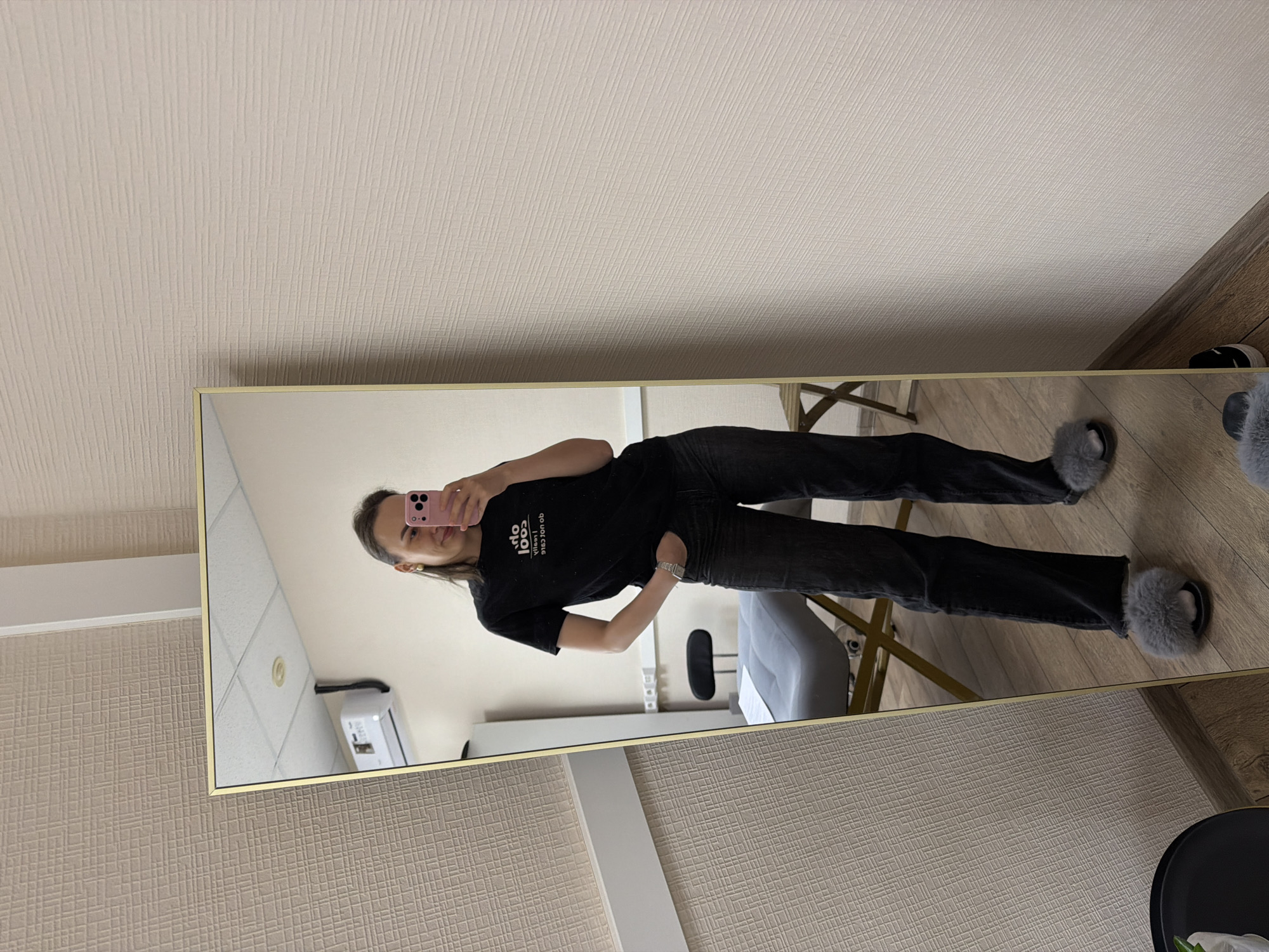 The image, presented rotated 90 degrees clockwise, captures a full-length mirror selfie of a woman.
**Subjects:** The central subject is a woman, standing casually and taking a picture with a pink smartphone held in her right hand. She has dark hair and a slight smile.
**Objects:** She is dressed in a black short-sleeved t-shirt, dark-wash jeans, and grey furry slippers. A silver-toned watch is visible on her left wrist. The mirror features a thin gold or brass-colored frame. The room outside the mirror has light beige textured wallpaper with subtle vertical lines and a crisscross pattern, and a light-colored wooden floor. A white baseboard runs along the wall. Partially visible objects on the floor include a dark, round table or stool and another grey furry slipper.
**Scene and Setting:** The setting is an indoor space, possibly a modern dressing room, waiting area, or a well-appointed room in a home or business in Chaikovsky, Russia.
**Activity:** The woman is posing for and taking a mirror selfie. Her left hand is casually placed in her jeans pocket.
**Notable Details:** The reflection in the mirror reveals a different view of the room: a light-colored wall, a ceiling with square panels and a circular light fixture, and a white air conditioning unit or similar appliance mounted on the wall. Part of a grey upholstered chair or stool is also reflected. The general lighting suggests it is daytime.
**Visible Text:** On the woman's black t-shirt, as reflected in the mirror, white text reads "oob yllacs cool." When read correctly (not reflected), this text spells "cool scallyboo."