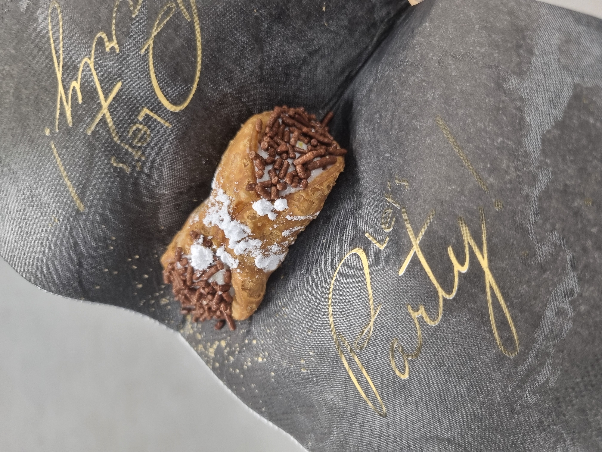 A single cannoli, a classic Italian pastry, is prominently featured in a close-up shot. The cannoli has a golden-brown, crispy shell, likely fried, and is filled with a white creamy substance, possibly ricotta cheese. Both ends of the cannoli are generously covered with small, dark brown chocolate sprinkles. A dusting of white powdered sugar is visible primarily on one side of the pastry shell and spilled onto the surface it rests on.
The cannoli is placed on a dark grey, possibly black, paper or fabric surface that has a subtle textured pattern. Gold-colored text is printed on this surface. The text clearly reads "Let's Party!" in a decorative, cursive font. The overall lighting suggests an indoor setting. The image focuses entirely on the dessert and its immediate surroundings, with no people visible. The context for this image is Saarbrücken, Germany.