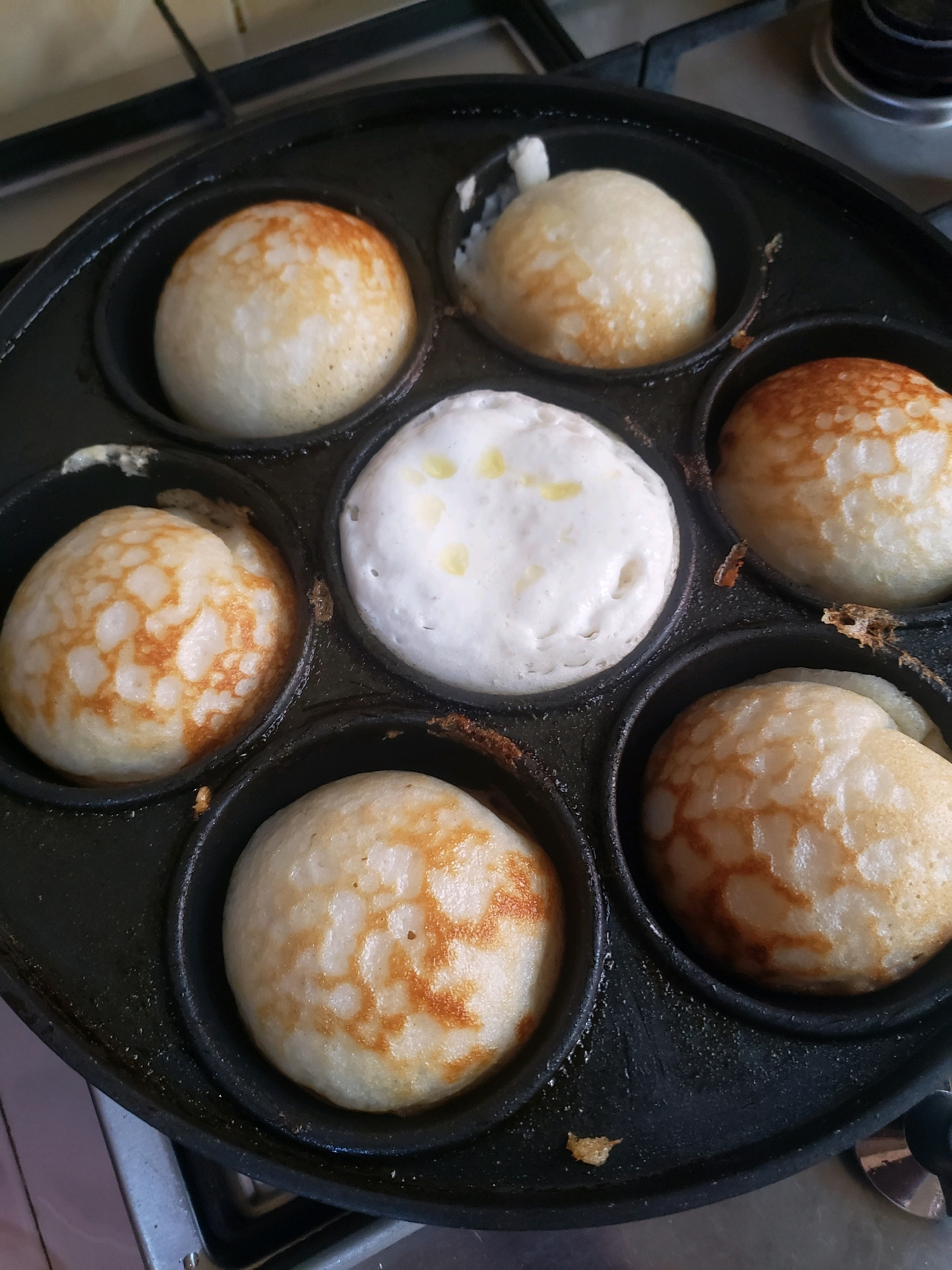 In Sabon Tasha, Nigeria, a black appam pan sits on a stove, containing seven small, round, puffed-up food items. Six of these items are golden brown with lighter brown, patterned markings, indicating they are cooked. One item in the center, however, remains pale white and appears to be uncooked batter, with a few droplets of oil visible on its surface. The appam pan has circular indentations where each food item is cooked. The background shows a portion of a kitchen stove.
