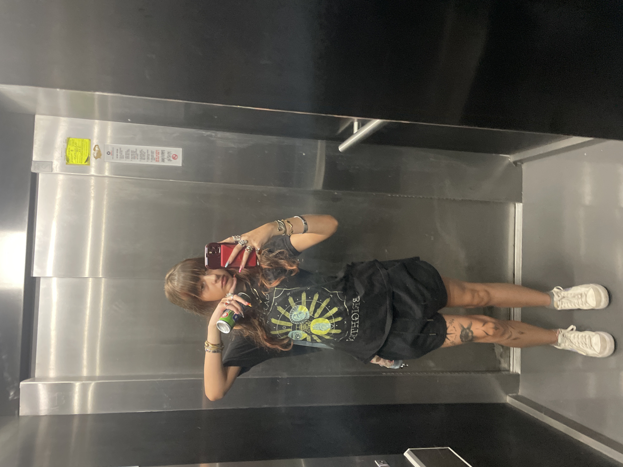A young woman with brown hair and bangs stands in an elevator, taking a mirror selfie with her phone. She is wearing a black t-shirt with a graphic print, black shorts, and white high-top sneakers. She has several rings on her fingers and bracelets on her wrists. She is holding a green can in her left hand. The elevator has stainless steel walls and a black panel on the right. There are some stickers on the wall to the left. The floor is gray. This image was taken in the city of Raanana, Israel.
