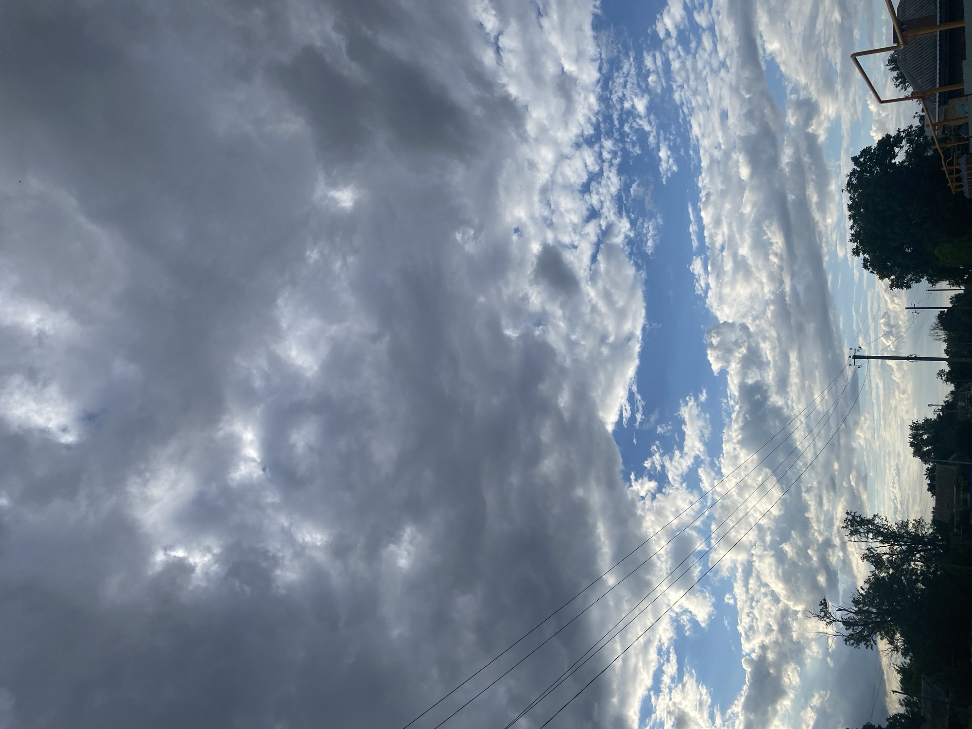 The media file is a photograph of a dynamic sky with varying cloud formations. The majority of the frame is filled with large, dark, and ominous storm clouds on the left, interspersed with patches of bright white and light grey cumulus clouds. To the right, the sky transitions to a clear blue, broken by streaks of smaller, wispy clouds that catch the sunlight, creating a dramatic contrast.
Visible in the lower right portion of the image are silhouetted trees, suggesting an outdoor setting with natural elements. A power line with multiple wires stretches diagonally across the frame, disappearing into the clouds and trees, hinting at a semi-urban or rural environment. A hint of a building structure with a yellow railing is visible at the top right edge, providing a further clue to the location.
The lighting suggests it is either early morning or late afternoon, with the sun's rays filtering through the clouds, creating shafts of light and casting shadows. The overall mood conveyed is one of dramatic weather, with the juxtaposition of dark storm clouds and bright, sunlit skies. There is no visible text in the image. Given the provided location context of Taganrog, Russia, the scene likely depicts the sky over that region.