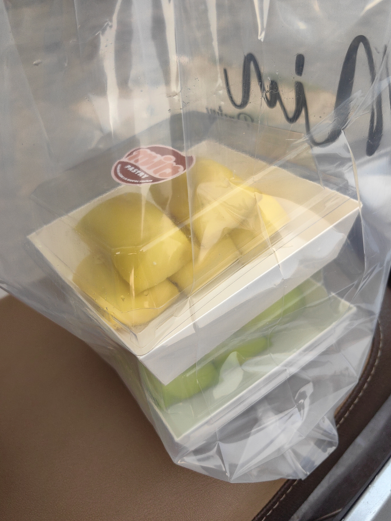 This image depicts two white, rectangular pastry boxes stacked inside a clear plastic bag. The top box contains yellow, potentially mango-flavored pastries, while the bottom box contains green pastries. A sticker with the word "pastry" is partially visible on the top box. There is writing in black ink, which appears to say "vaj," on the plastic bag. The scene is located in the city of Ha Lam Ward, Vietnam. The bag containing the pastries is resting on a brown surface.
