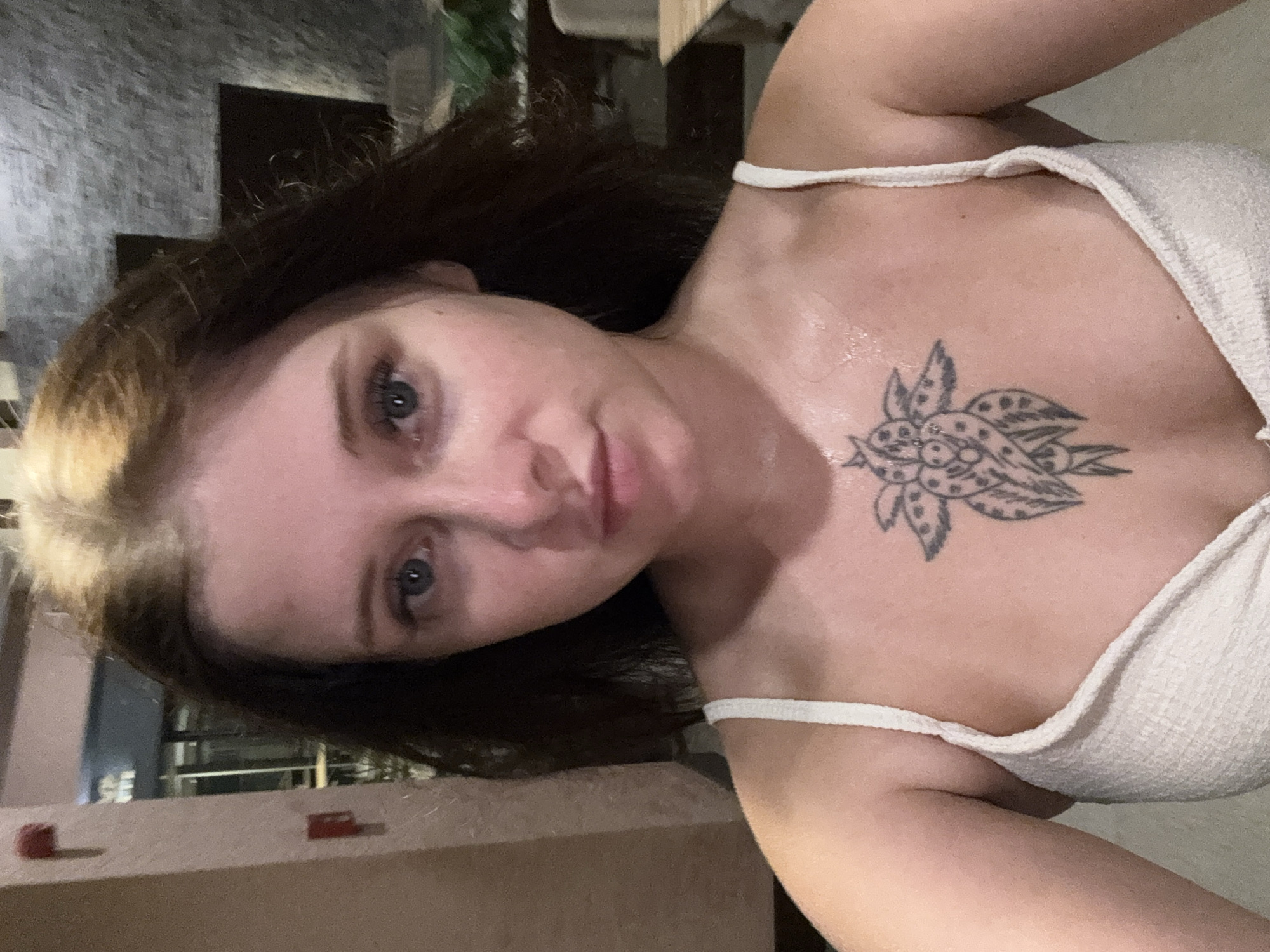 A medium shot shows a young woman with light skin and brown hair pulled back from her face, looking directly at the camera. She is wearing a white textured tank top, revealing a tattoo on her chest that resembles a stylized flower or sun with a stem and leaves. The background is dimly lit, with a textured wall to the left and what appears to be a dark stone or brick wall behind her. To the right, a blurred glimpse of greenery and furniture suggests an indoor or covered outdoor setting, possibly a restaurant or lounge area. The woman’s expression is neutral, and her skin appears slightly dewy, suggesting a warm environment or recent activity. The lighting is soft, with the woman's face being the primary illuminated subject.