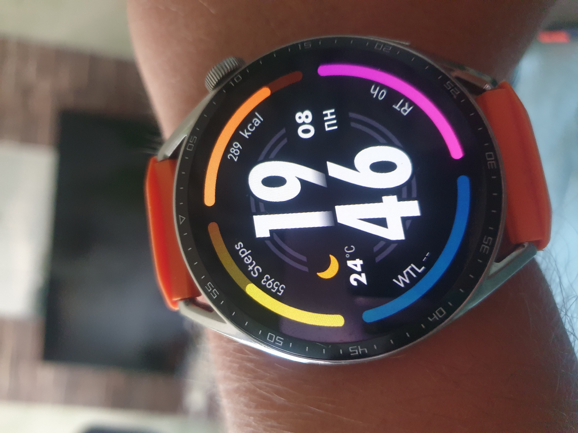 The image displays a close-up of a smartwatch worn on a person's wrist. The smartwatch has a dark circular face with various information displayed. Prominently featured are the time "19 46", the temperature "24°C" with a crescent moon icon indicating it is nighttime, "5593 Steps", and "289 kcal". There are also segments of colored progress rings and some text which appears to be "WTL--" and "RT 0h". The watch has an orange strap. The background is blurred, showing what appears to be an indoor setting with a dark rectangular object (possibly a television) and a textured wall behind it. No specific location in Sofia, Bulgaria, can be identified from the image.