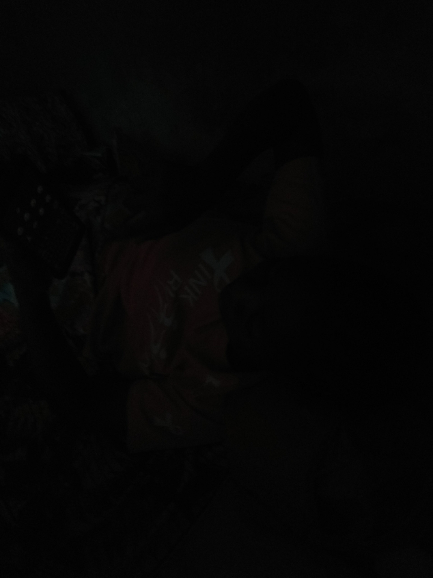 The image depicts a dark room with a person lying down. The person is wearing a pink shirt with white lettering that appears to spell out "PINK FLOYD." The person is on their side, facing away from the camera. To the left of the person, a portion of what looks like a mobile phone or a remote control is visible, with several circular buttons. The background is very dark and indistinct, making it difficult to discern the environment beyond the immediate vicinity of the person and the device. The overall lighting is extremely low, suggesting a dimly lit or nighttime setting. There are no clear indicators of the specific location within Jalingo, Nigeria, as the image lacks any recognizable landmarks or contextual elements that would pinpoint its geographical setting.