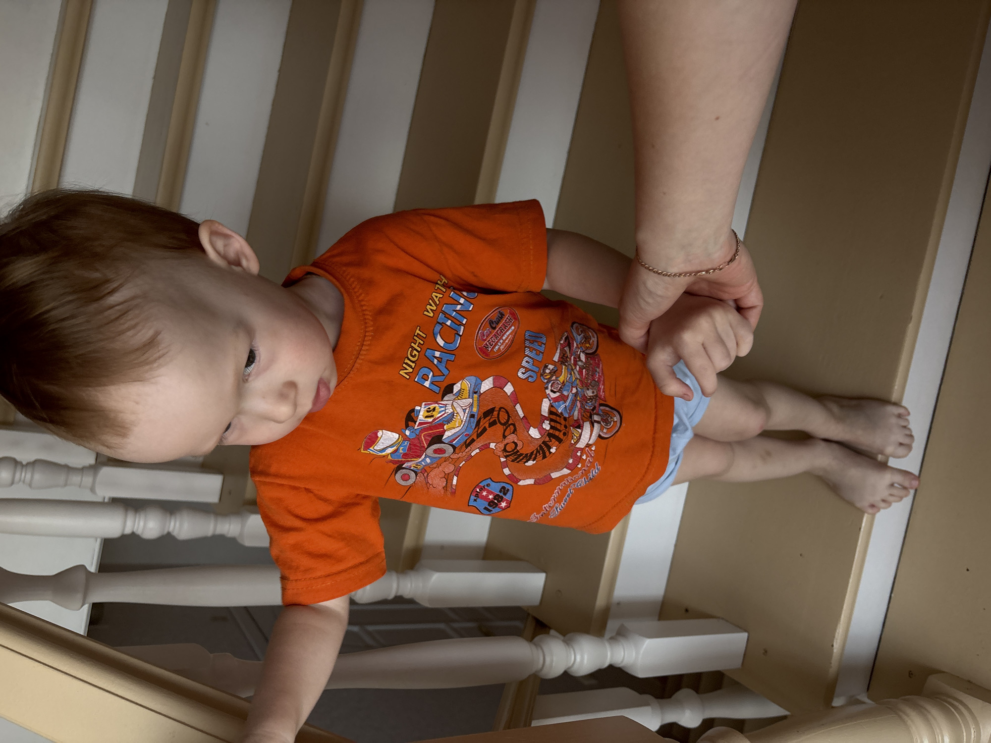 A child wearing an orange t-shirt with a racing motif and light blue underwear is standing on a staircase with white balusters and beige risers/treads. The child is holding onto the railing with one hand and has the other hand held by an adult. The child is looking down at the stairs. The location is unspecified, but given in the prompt as being in Ферма, Russia.