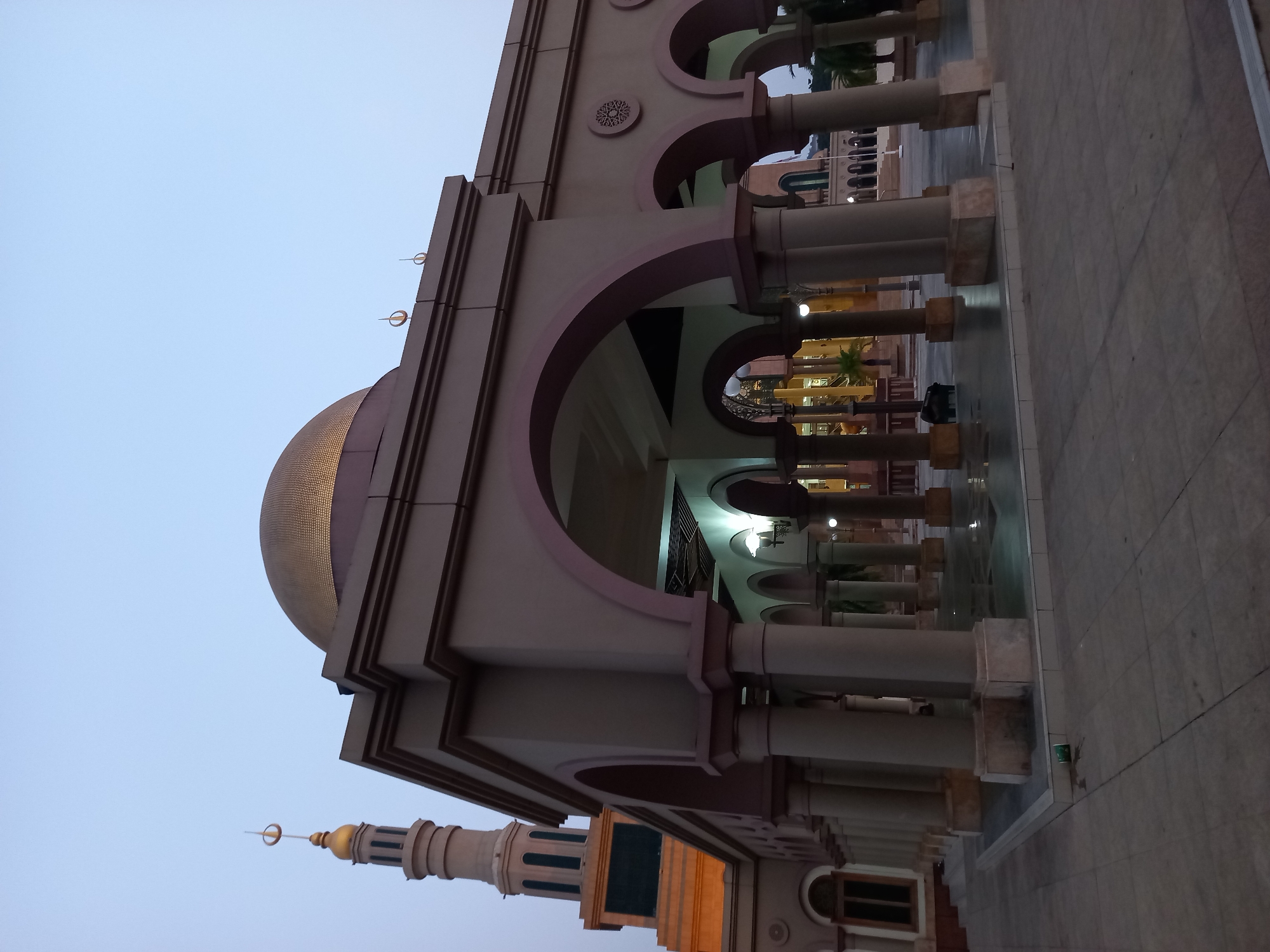 The image shows the exterior of a mosque with a golden dome and a tall minaret. The mosque has a colonnade with arched walkways in the foreground. The sky is a pale blue, suggesting it is either dawn or dusk. The ground in front of the mosque is paved with square tiles. A single green cup is visible on the ground near the base of one of the pillars. The architectural style is consistent with Islamic religious buildings. This mosque is likely the Islamic Center Samarinda, also known as Masjid Islamic Center Samarinda, located in Samarinda, East Kalimantan, Indonesia.