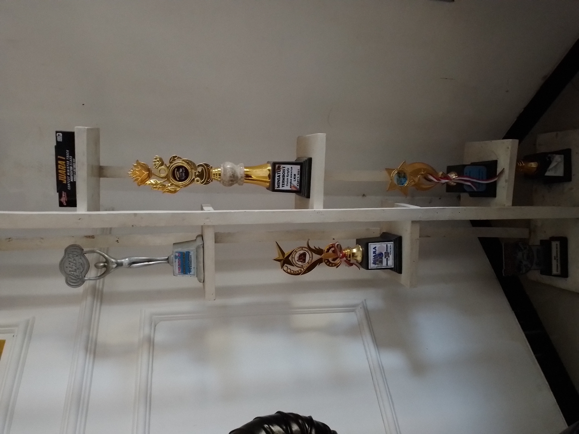 The image displays a white, multi-tiered shelf mounted on a wall in a dimly lit room. The shelf holds several trophies and what appears to be a plaque. On the top shelf, a silver trophy with a circular emblem at the top is visible. Below it, on the next shelf, is a black plaque that reads "JUARA 1 CONTEST MINIATUR TRUCK MODIFICATION CLASS". On the third shelf, a golden trophy with intricate designs and a star at the top is placed. This trophy has a black base with text that includes "MINIATUR TERMOBOIS 1". The bottom shelf holds another trophy, mostly obscured by shadow, with a black base. The shelves are made of a light-colored material, possibly wood or laminate, and are supported by a white vertical structure. The wall behind the shelves is painted white and has visible trim details. The overall impression is that of an indoor display area, possibly a home or office, in Ngancar, Indonesia.