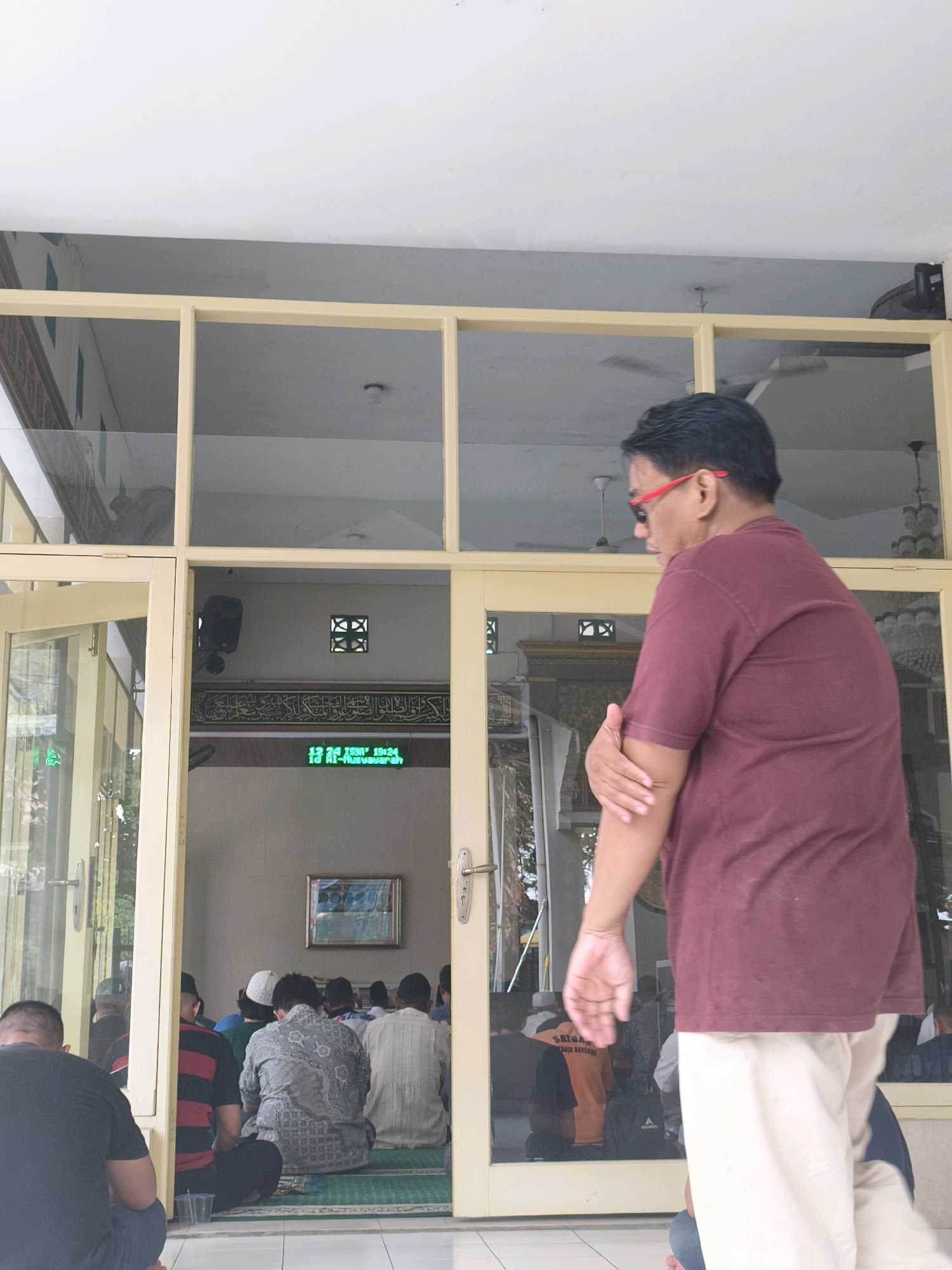 A group of men are gathered inside a mosque in Samarinda, Indonesia, facing towards the front of the room, seated on a green carpet. A man in the foreground, wearing a maroon t-shirt and light-colored pants, is standing and looking towards the mosque interior. The mosque has glass doors and windows, allowing a view of the interior. A digital clock displaying time and prayer information is visible on the wall inside the mosque.