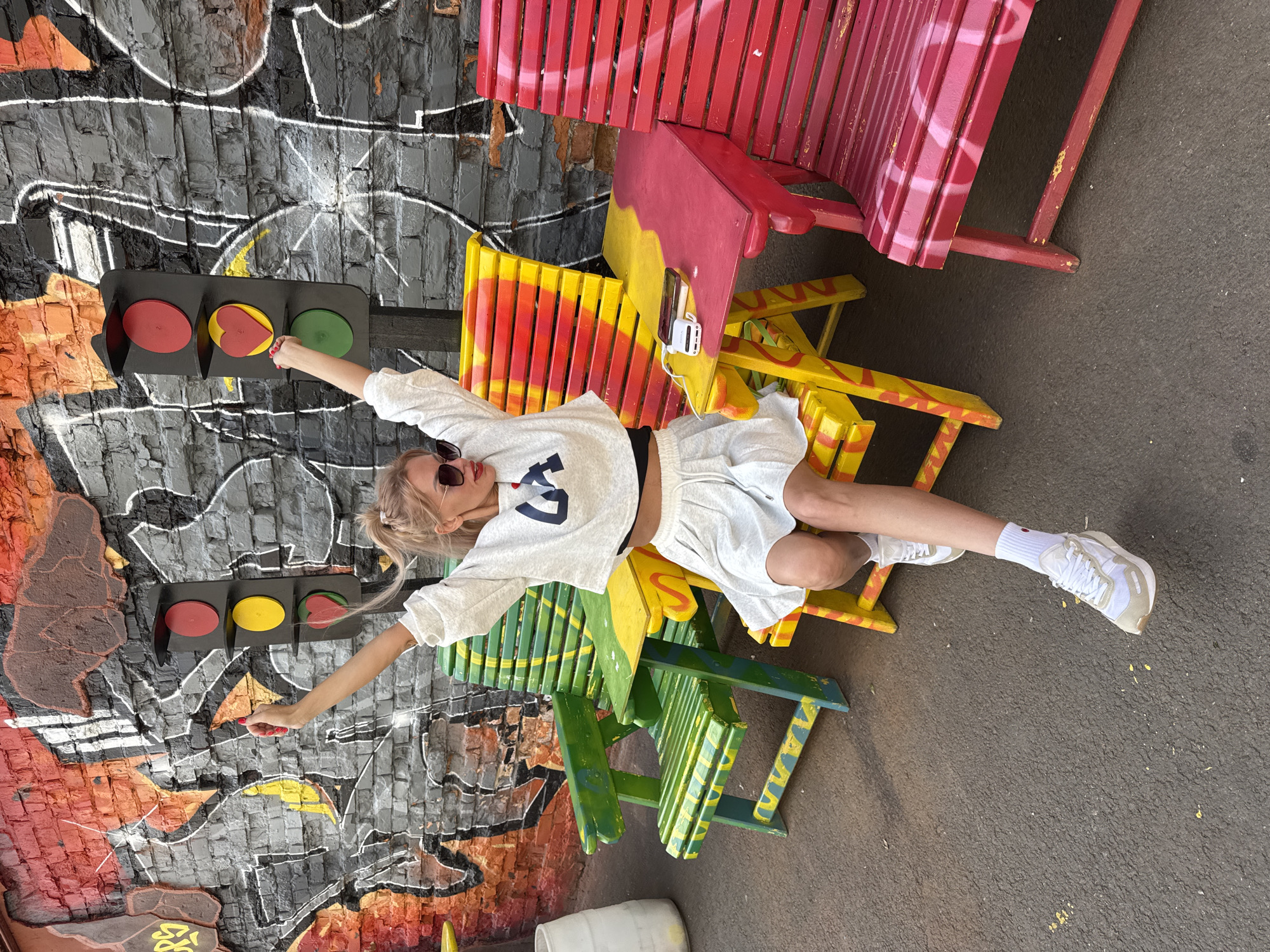 In Saint Petersburg, Russia, a young Caucasian woman with blonde hair tied in a ponytail, wearing sunglasses and a white cropped sweatshirt with dark lettering and a white pleated skirt, sits on a brightly colored yellow and red bench. Her arms are raised in a gesture of excitement, and her right hand is reaching towards a traffic light that has been creatively modified with a red heart in the center instead of the traditional yellow light. The background features a brick wall adorned with vibrant graffiti, creating a playful and urban aesthetic. Several other colorful benches, painted in shades of green, yellow, red, and pink, are visible in the scene, along with a small table on which a mobile phone and a white portable charger are placed. The ground is paved with asphalt.