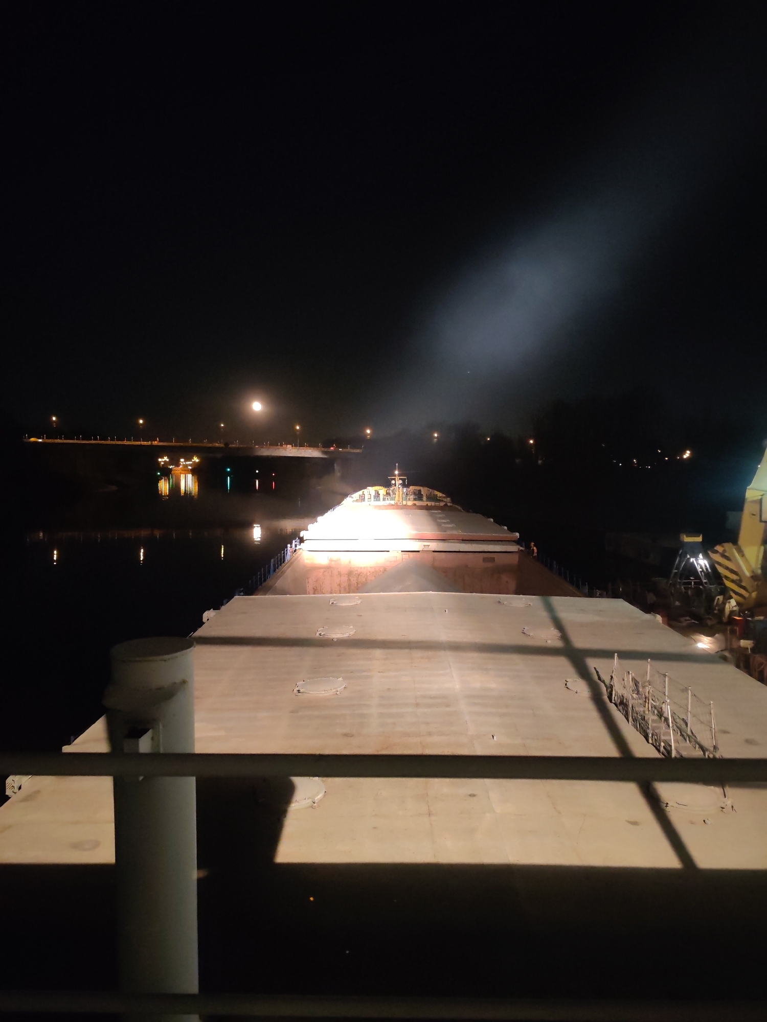 The image shows a night scene, likely in Yakhroma, Russia, given the prompt. A barge dominates the foreground, its flat surface illuminated by onboard lights. A low metal railing and white cylindrical post are visible in the immediate foreground, suggesting the viewpoint is from another part of the barge or a nearby structure. Beyond the barge, a bridge spans the water, with streetlights illuminating its length and reflections visible in the water below. A bright moon is positioned near the bridge. In the background, on the far side of the water, are trees and buildings, also illuminated with lights. A beam of light from an unknown source cuts through the air, adding to the overall illumination. There is no indication of people other than shadows which could indicate them. A crane, potentially for loading or unloading the barge, is seen on the right of the image, close to the boat.