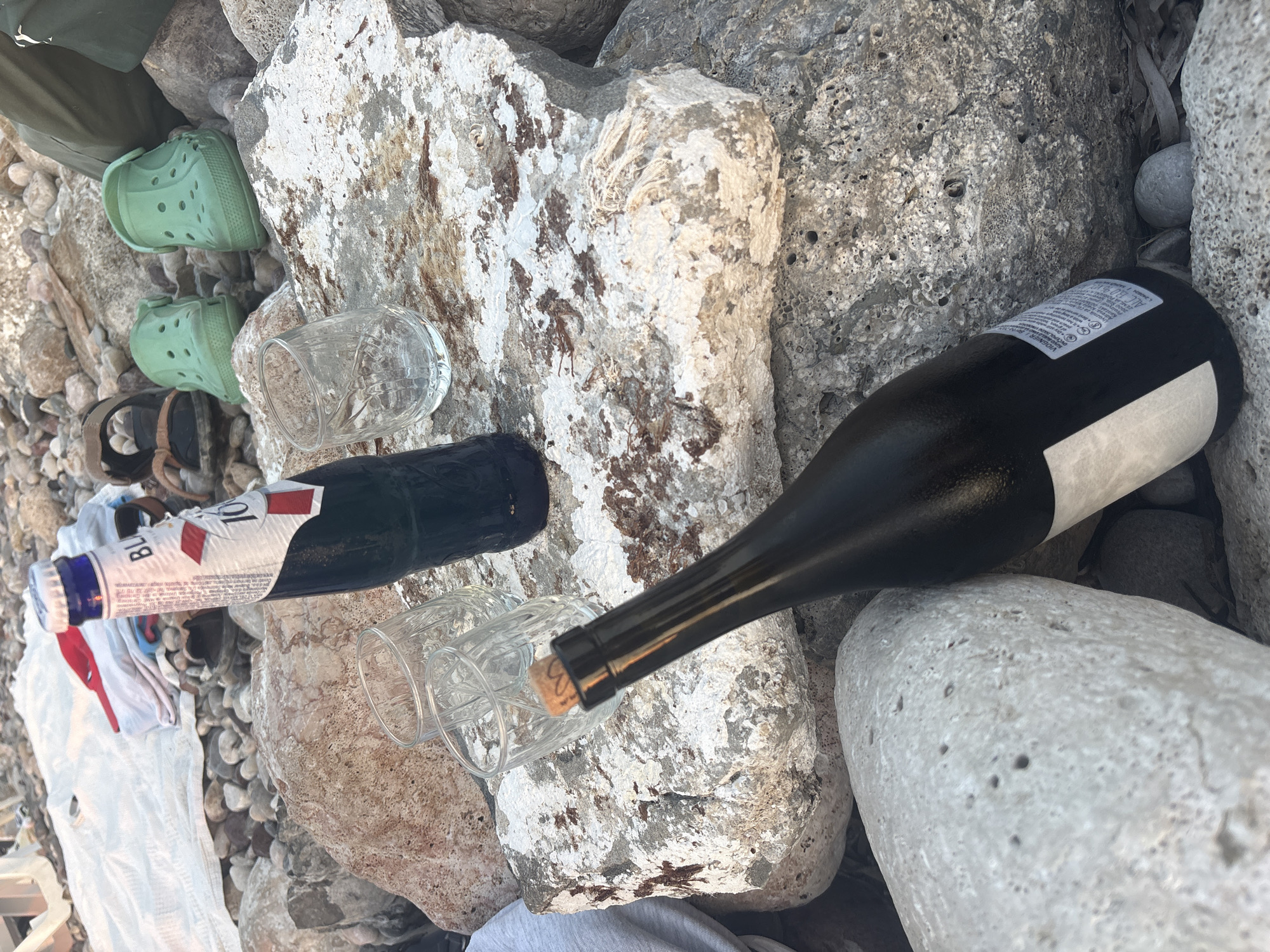 The image depicts an outdoor scene on a rocky shore, likely in Bar, Montenegro. In the foreground, a dark glass bottle with a cork stopper is angled towards the camera. To its right, another dark glass bottle with a white label, partially obscured by the first bottle, is visible. Two empty glass tumblers are placed on a large, light-colored rock in the middle ground. Behind the tumblers, a blue-capped bottle with a white label displaying red accents and text is standing upright. Scattered around the bottles and glasses are various rocks of different sizes and colors, some appearing porous and weathered. In the background, a white fabric is spread out on the rocks, with some folded clothing items and a pair of dark sandals resting on it. Further to the right, two bright green clogs are visible, and parts of what appear to be green fabric pants suggest the presence of at least one person partially out of frame. The overall setting suggests a casual gathering or picnic by the water, characterized by natural elements and recreational items.
