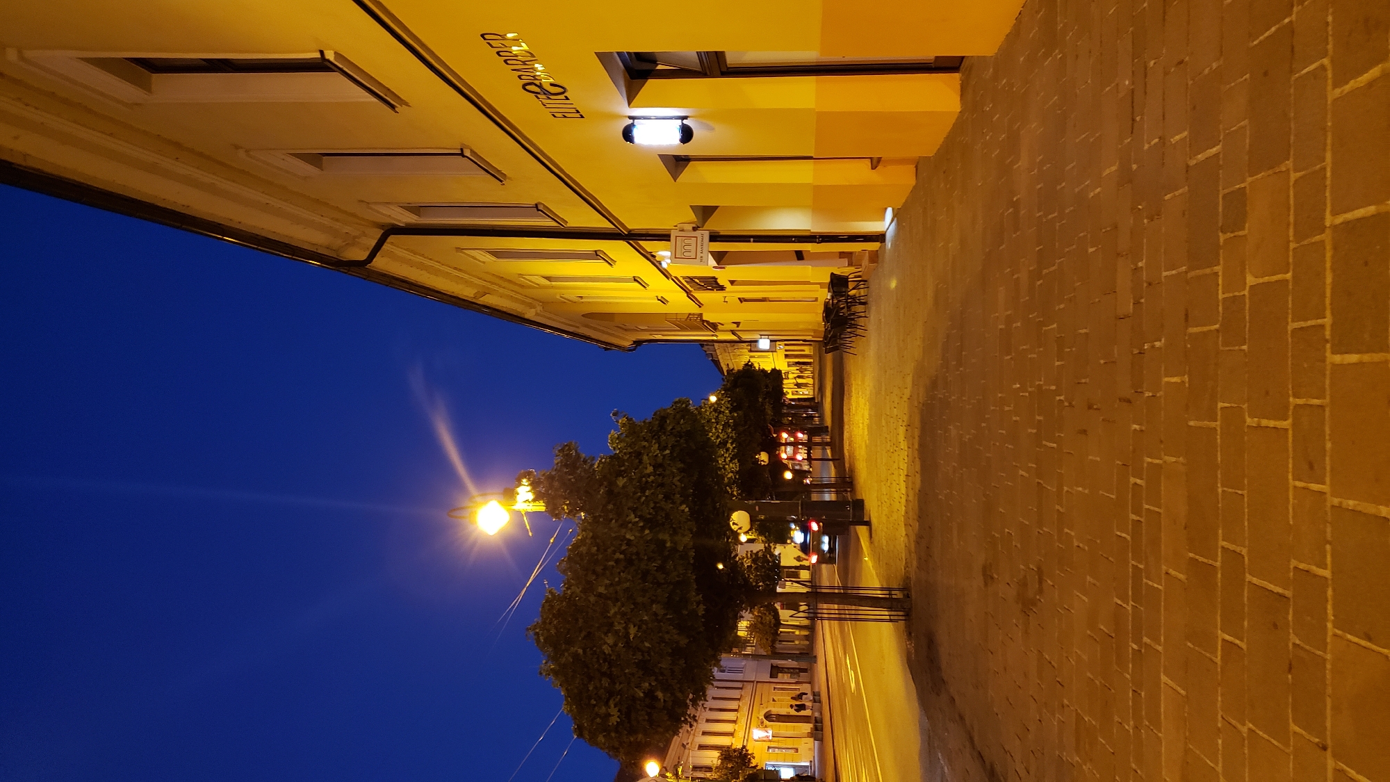 The image depicts a street scene in Prešov, Slovakia, during twilight. The sky is a deep blue, transitioning from daylight. On the right side of the frame, a long, yellow building lines the street, its facade illuminated by streetlights and interior lighting. A sign reading "ELITE & BARBER" is visible on the building. The sidewalk in front of the building is paved with light-colored bricks. Several outdoor tables and chairs are arranged on the sidewalk. On the left side of the street, a row of trees with full canopies are planted along the sidewalk. Beyond the trees, a street with traffic and other buildings is visible. A streetlight with a bright yellow bulb illuminates the scene. The overall atmosphere is calm and quiet, characteristic of an evening in a city.