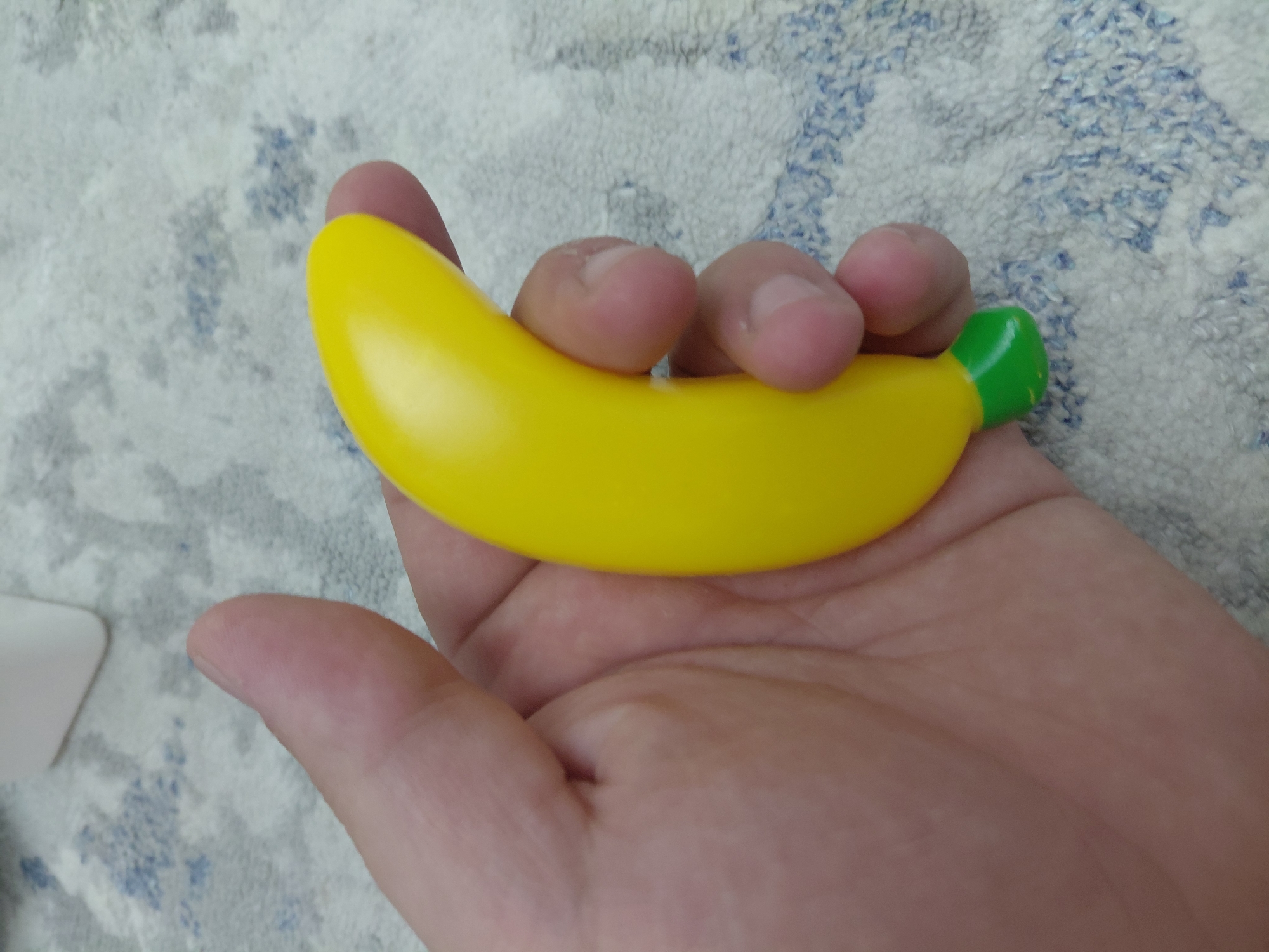 A hand is holding a yellow banana-shaped toy with a green tip. The hand is positioned in the foreground, with the toy cradled in its palm and fingers. The background shows a patterned surface that appears to be a rug or fabric. The lighting suggests an indoor environment. There is no indication of the city of Aktau, Kazakhstan in the image.
