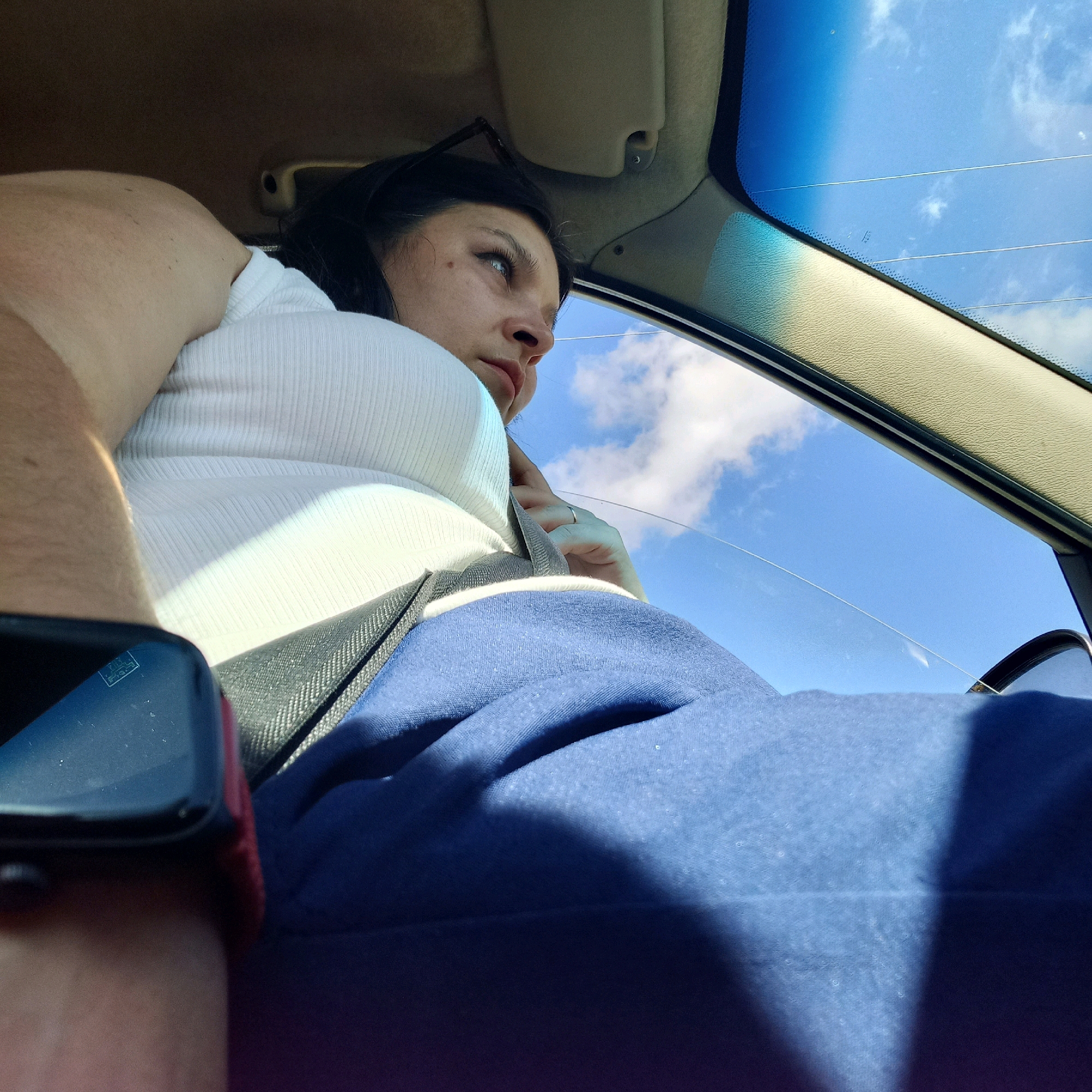 A woman in a white ribbed top and blue sweatpants is sitting in a car, looking out the window. She has dark hair and is wearing a watch on her left wrist. The car's interior, including the headliner and sun visor, is visible. The window shows a bright blue sky with scattered white clouds. The image is taken from inside the car, looking out. There is no information about the location being Klyntsi, Ukraine.