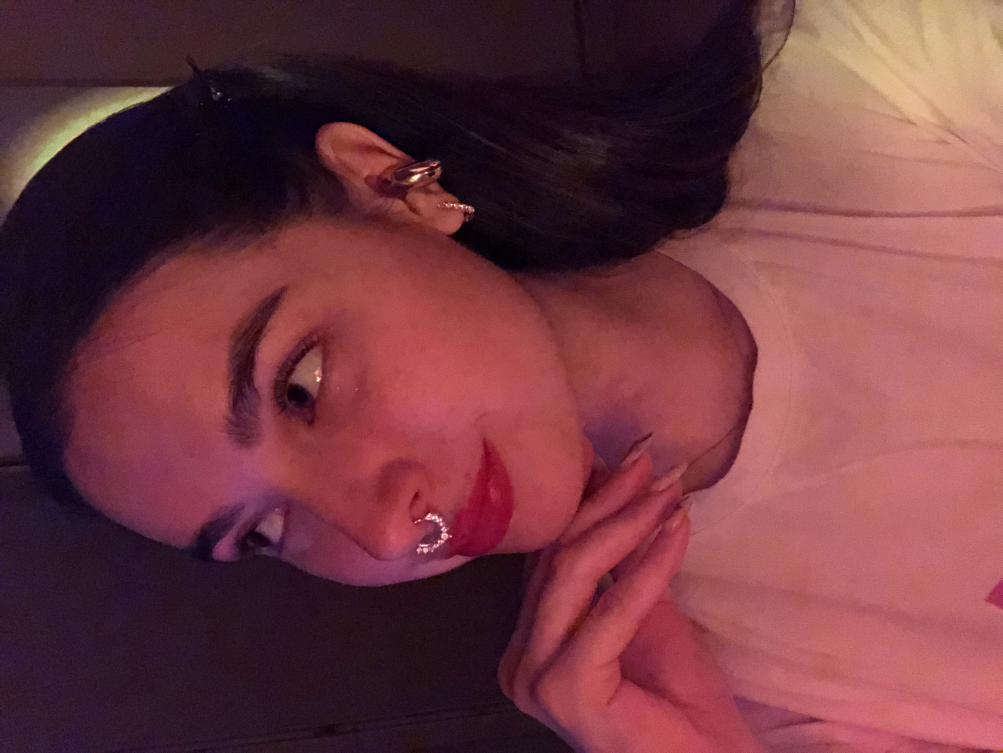 A woman with long, dark hair is seen in an indoor setting. She is wearing multiple earrings on her left ear. She has a small ring in her nose. Her hands are near her face. She is wearing a white shirt. The location cannot be determined.