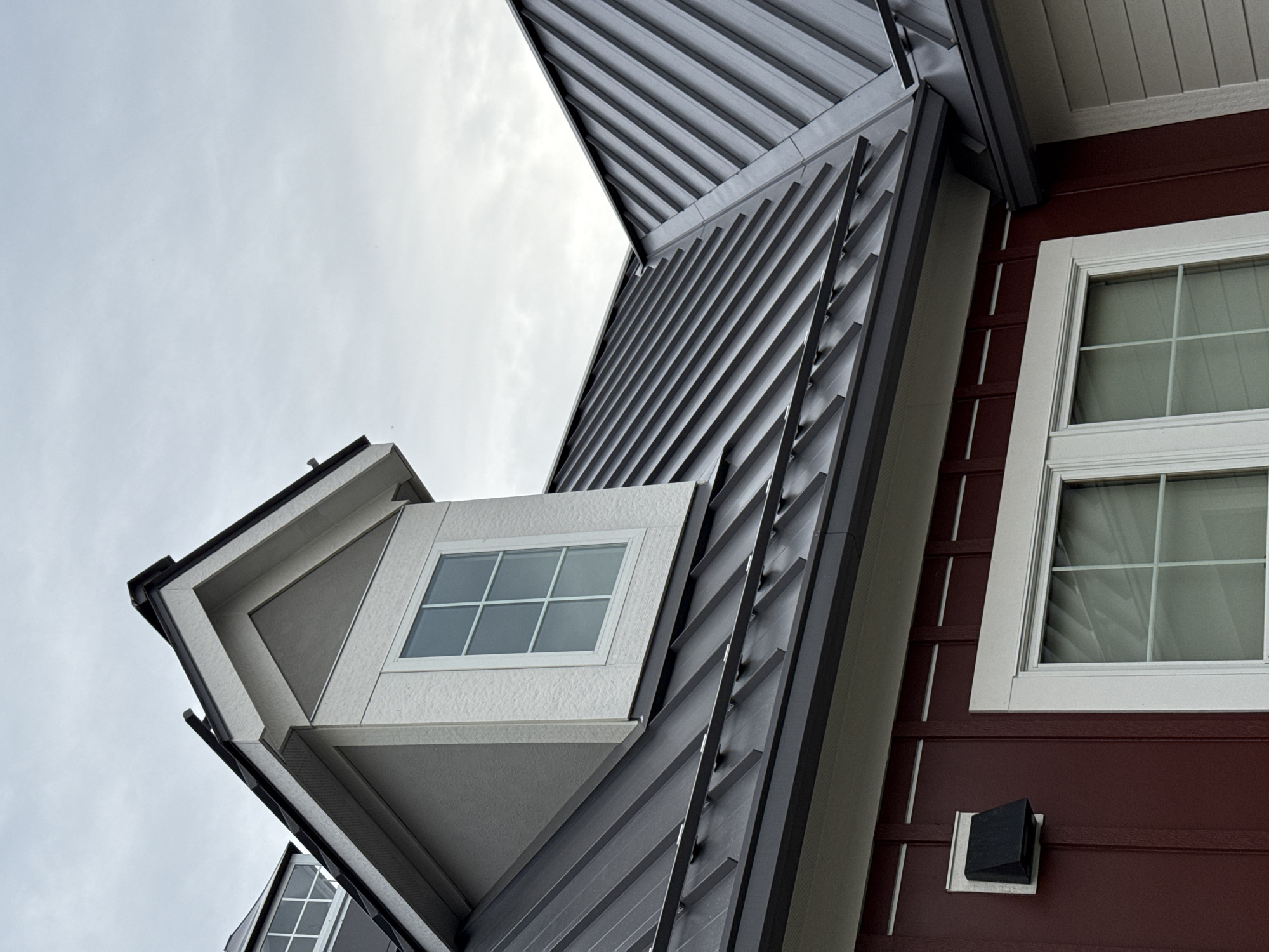 A low-angle shot captures a unique architectural design against a cloudy sky. The building features a combination of materials, including dark gray metal siding with linear accents and maroon-colored horizontal siding. Several windows with white frames are visible, one extending outwards from the structure at an oblique angle. The building's design incorporates geometric shapes and varying textures, adding to its contemporary aesthetic. The location is identifiable as Grove City, United States of America. No people are visible in the shot.