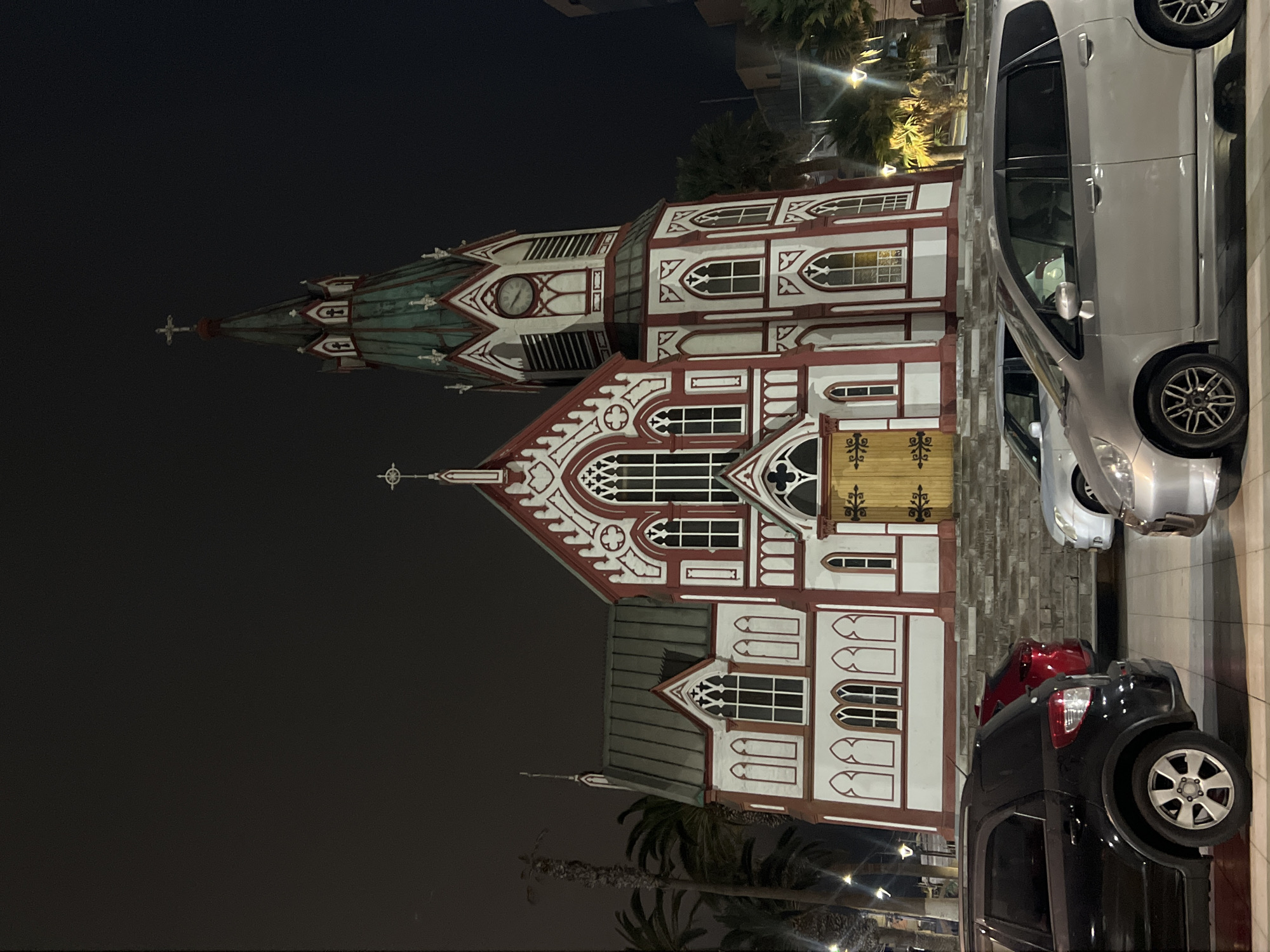 The media file captures a nighttime view of the Catedral de San Marcos de Arica, Chile, a distinctive wooden Neo-Gothic church. The building is prominently illuminated, showcasing its white walls, intricate red trim, and green/teal spire.
The central subject is the ornate church, characterized by its tall, slender spire topped with a cross. A clock face is visible on a section of the spire. The main body of the church features multiple pointed-arch windows and rectangular windows with decorative tracery, all framed by elaborate red and white patterns. A notable wooden double door with dark metal decorative elements is visible towards the front of the structure. The architecture suggests a meticulous wooden construction, likely prefabricated.
In the foreground and to the right, several cars are parked along a street. A silver hatchback, a red car (partially obscured), and a black SUV are clearly visible, with streetlights reflecting off their surfaces. Palm trees, also illuminated by artificial light, dot the background, suggesting a tropical or subtropical climate. The ground includes paved road and stone-textured sidewalk areas.
The scene takes place at night, with the dark sky contrasting sharply with the brightly lit building and street. Artificial lights from the building's interior and surrounding streetlights cast a warm glow over the scene. No specific activity or event is taking place, presenting a static streetscape. While a clock face is visible on the spire, no discernible text or numbers can be read from it. The overall impression is one of architectural beauty set against the night sky in an urban environment.