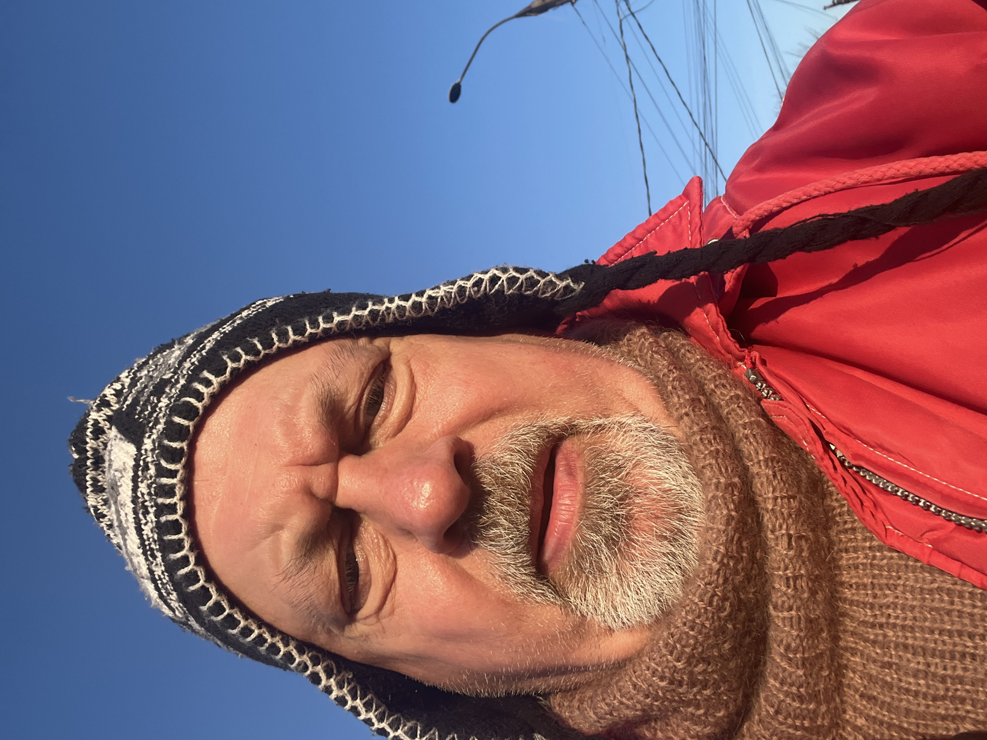 A low-angle selfie features an adult male wearing a red jacket with a black drawstring hood, a black and white knit hat, and a brown knit scarf. The man has a light complexion, graying beard and mustache, and his eyes are squinting. Behind him, a clear blue sky is visible, with a street lamp and power lines also in view. It is not possible to confirm that the location is in the city of Podolsk, Russia, from the visual information provided.