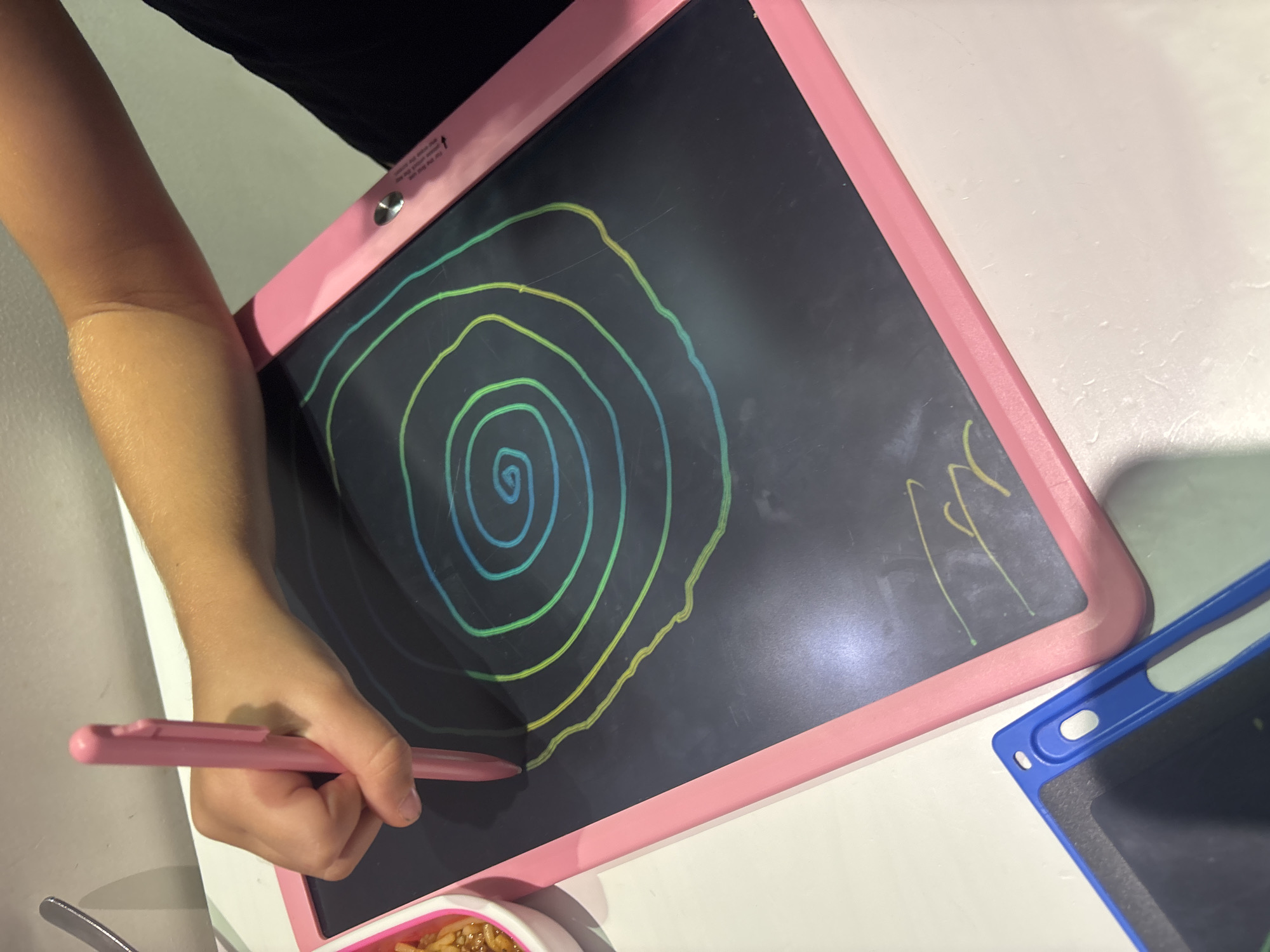 A child's hand holds a pink stylus, drawing a colorful spiral on a black LCD drawing tablet. The tablet has a pink border. Another blue tablet is partially visible in the foreground. The scene is indoors, likely on a table. The presence of food in a white bowl to the left suggests a casual setting, possibly a home or cafe. There is no information to suggest this location is in Dubai, United Arab Emirates.