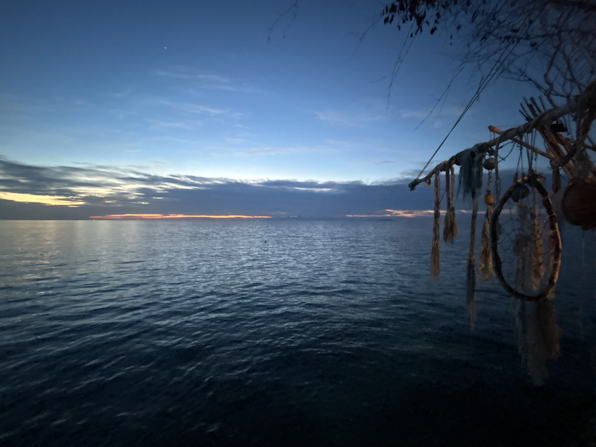 The image depicts a serene twilight scene over the ocean, likely on Koh Phangan, Thailand. The sky is a gradient of deep blue at the top, transitioning to lighter blues and then to a pale orange and yellow band near the horizon, indicating the afterglow of sunset. Wispy clouds are scattered across the sky. The ocean is calm, with gentle ripples reflecting the sky's colors. In the foreground, on the right side of the frame, a collection of dreamcatchers and other hanging decorative items are suspended from what appears to be a wooden structure or branch. These items are rendered in silhouette and partially obscured by darkness. The overall atmosphere is peaceful and tranquil, with a hint of bohemian decor.