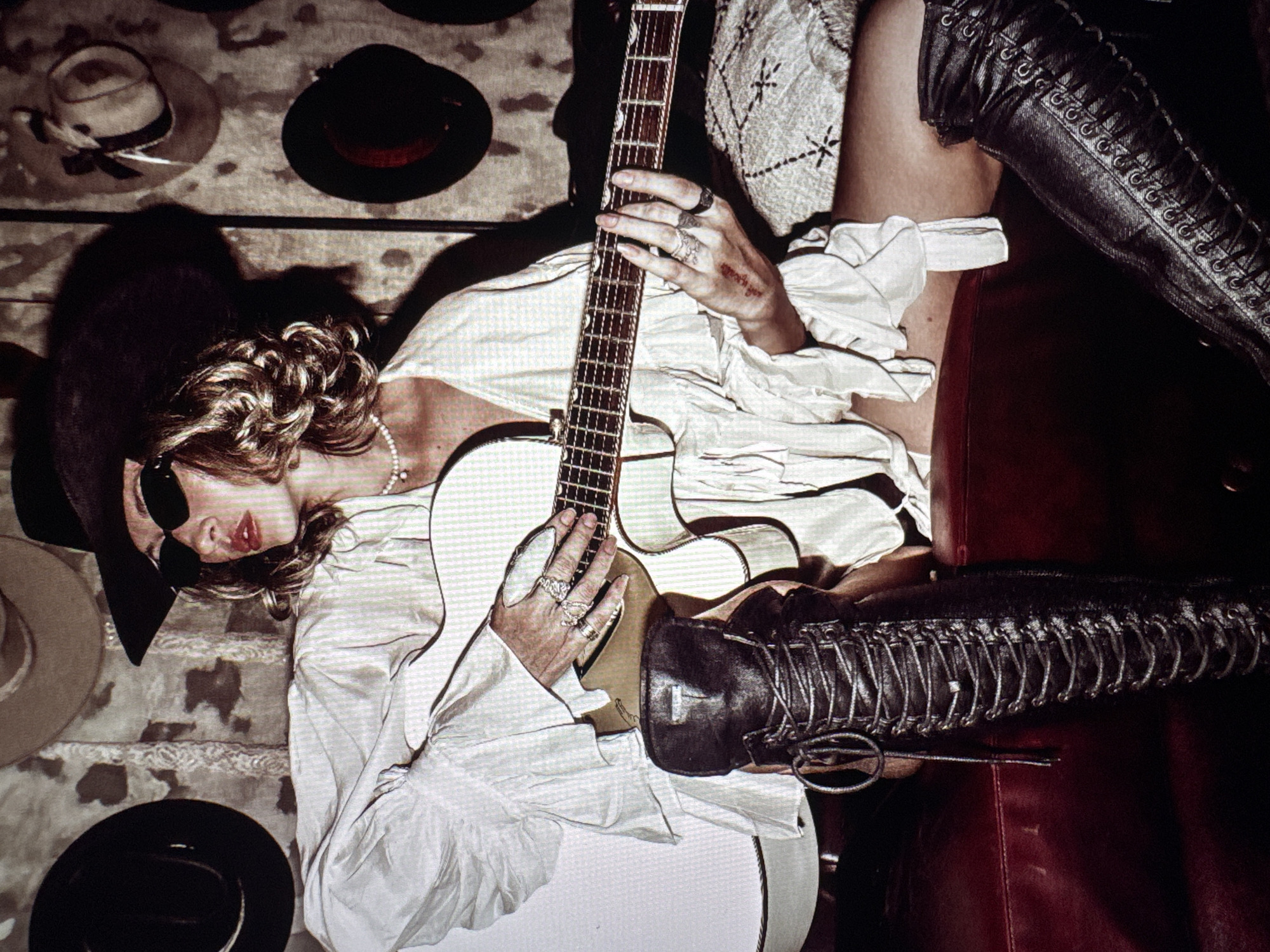 A woman wearing a white blouse, black hat, sunglasses, a pearl necklace, and lace-up black boots is lying down and holding a white electric guitar. Several hats hang on the wall behind her. The location, Mengwi, Indonesia, cannot be verified from the visual information provided.