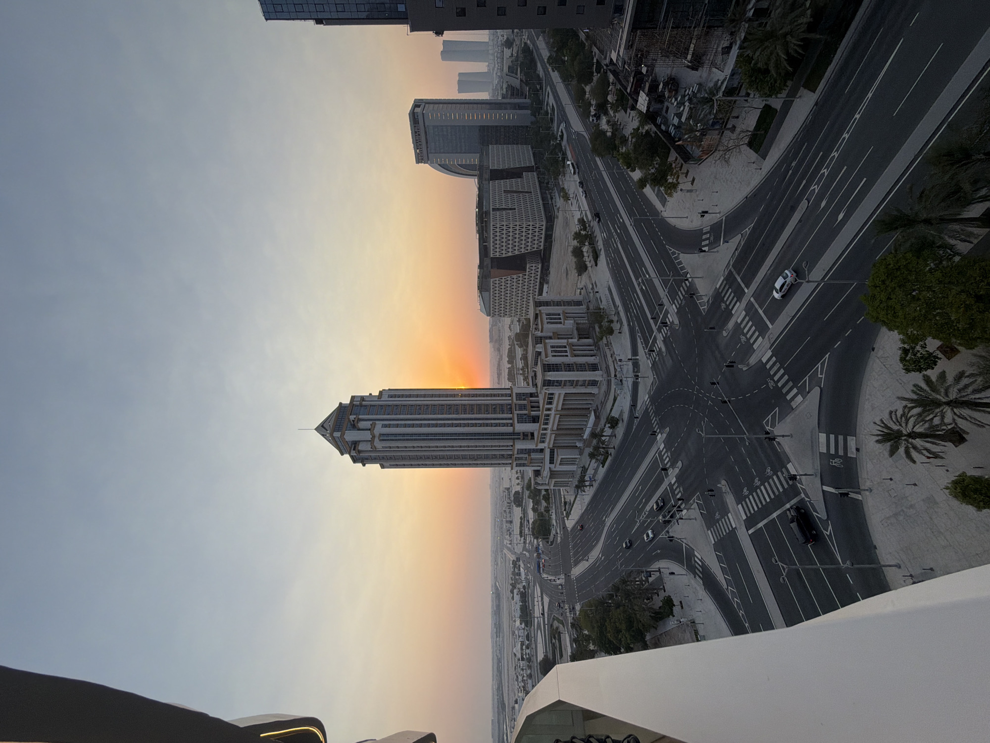 The image captures an aerial view of a modern urban intersection in Lusail, Qatar, during sunset. A tall, distinctive skyscraper with a pointed roof dominates the right side of the frame. Several other contemporary buildings, including a curved architectural structure, are visible in the background. The intersection itself is wide and clearly marked with traffic lanes and pedestrian crossings. A few vehicles, including cars and a dark SUV, are navigating the roads. The sky displays a gradient of soft orange and yellow hues from the setting sun, contrasting with the clear blue sky above. Palm trees line some of the sidewalks and medians, indicating a warm climate. The overall scene depicts a developed city with a mix of residential and commercial architecture, bathed in the warm glow of dusk.