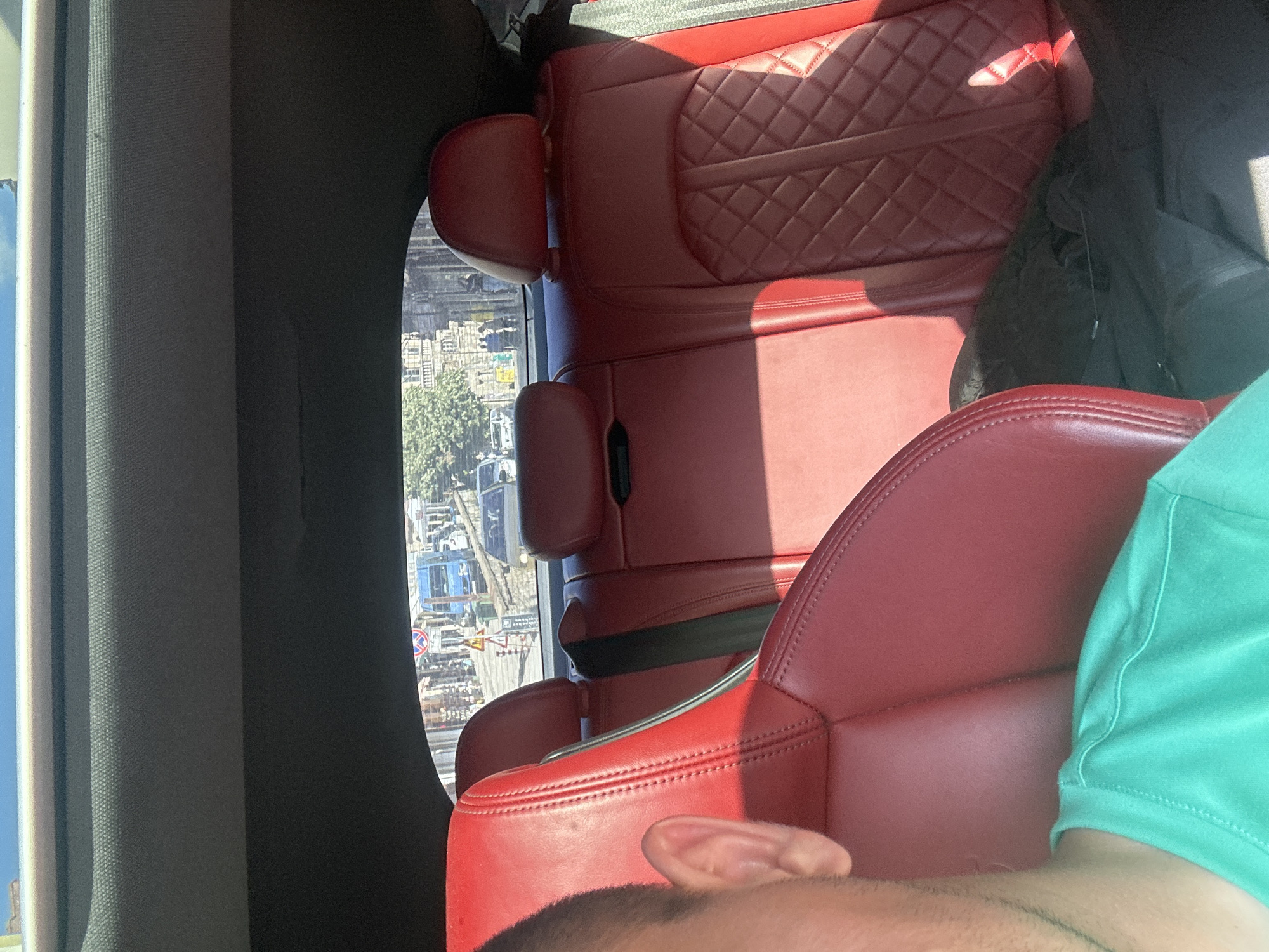 The image captures a detailed view from inside a car, looking out through the rear window onto an urban street scene in Veliko Tarnovo, Bulgaria.
In the foreground, the luxurious red leather interior of the car is prominent. On the far left, the back of a front passenger seat is visible, featuring vertical stitching. The rear left passenger seat's headrest and part of its backrest are also seen. To the right, the rear right passenger seat displays a distinctive quilted or diamond-stitched pattern on its backrest. A black object, possibly a bag or jacket, rests on this seat. The car's grey headliner is visible across the top. Partially visible on the far left, in the immediate foreground, is the left ear and shoulder of a person wearing a teal t-shirt, suggesting the photo was taken from the driver's side or left-hand side of the vehicle.
Through the rear window, a sunny street scene in Veliko Tarnovo unfolds. The characteristic architecture of multi-story stone buildings, typical of the city's historic center, lines both sides of the street. The road appears to have a slight downhill gradient. In the mid-ground, a light blue urban bus or large van is prominently positioned, slightly to the left of the frame's center. To its right, a white sedan is visible, with other vehicles further down the street. Distinctive road signs are visible: a red and white circular sign (likely "No Entry for all vehicles") is above and slightly to the left of the blue bus, mounted on a pole. Below it, a yellow triangular warning sign with an unclear black symbol is also present. Several pedestrians are scattered along the sidewalks and potentially crossing the street. Green foliage from trees is visible on the left side, partially obscuring some buildings. The bright sunlight casts strong highlights and shadows throughout the scene, both inside the car and on the street outside. The combination of the specific building styles, the urban bus, and the road signage points to a location within the historical or commercial areas of Veliko Tarnovo, such as ul. Stefan Stambolov or ul. Nikola Piccolo, viewed from a car likely positioned uphill or at a junction looking down one of these thoroughfares.