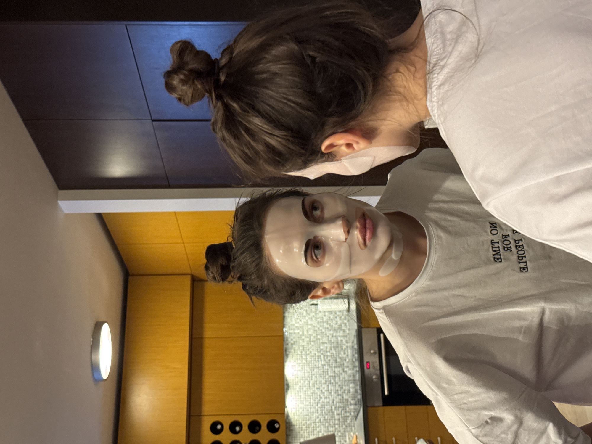 Two women wearing white sheet masks are indoors. One woman is looking at the camera. Both women are wearing white t-shirts and have their hair in buns. Behind them is a kitchen with wood cabinets, a wine rack, and a gray tiled wall. The location is Unknown, Unknown.