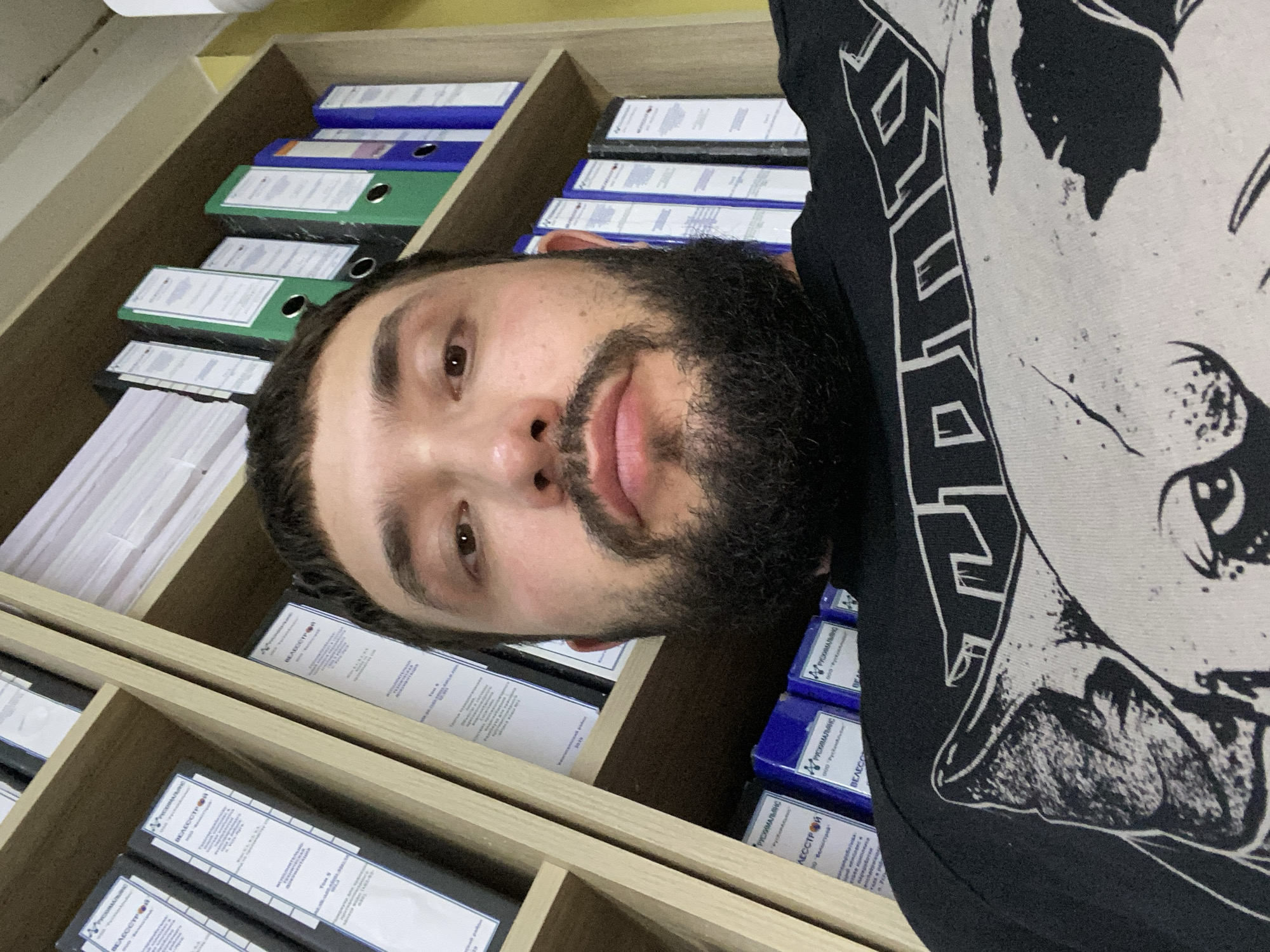 A close-up, low-angle, selfie-style photograph captures a man with a beard, positioned in front of bookshelves filled with binders. The man is wearing a black t-shirt with a graphic print. The environment appears to be an office or study, with the bookshelves forming the backdrop. The man's face, partially obscured by his beard, is the central focus. The binders on the shelves are of various colors, including white, blue, and green, and some have labels with Russian text. The lighting suggests an indoor setting.
