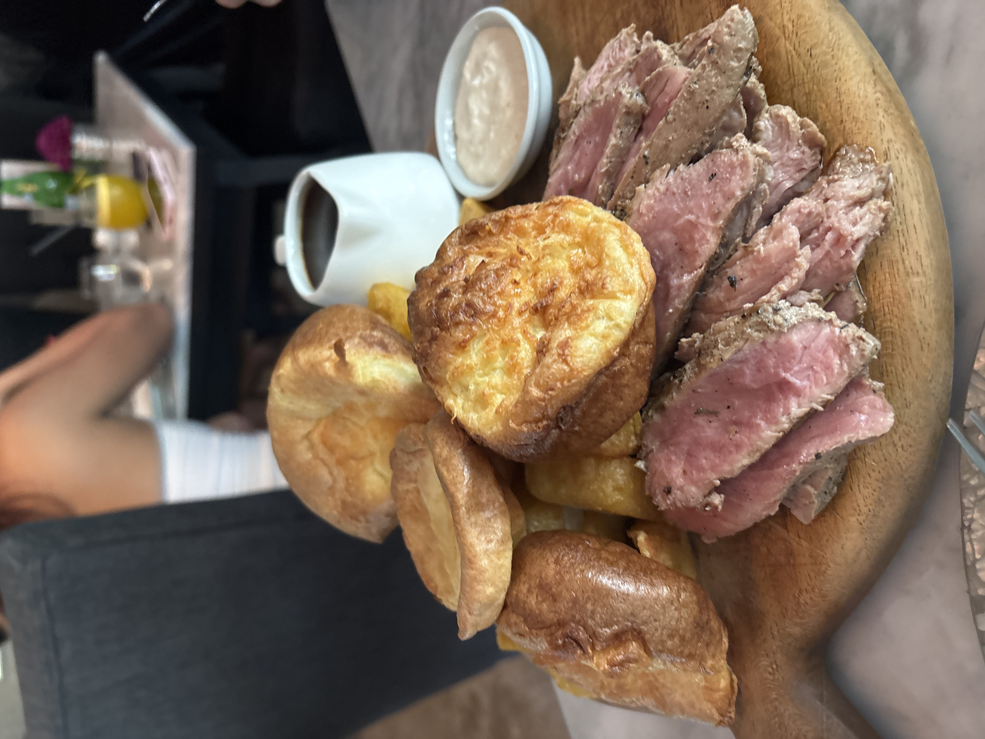 A wooden serving board is pictured, containing sliced rare roast beef, Yorkshire puddings, potato chips (fries), a gravy boat filled with dark sauce, and a small bowl of white sauce. A person's arm and gray clothing are visible on the left side of the image. A blurry background suggests a restaurant or cafe setting. The location of North Kuta, Indonesia is specified but not visually verifiable in the photograph.