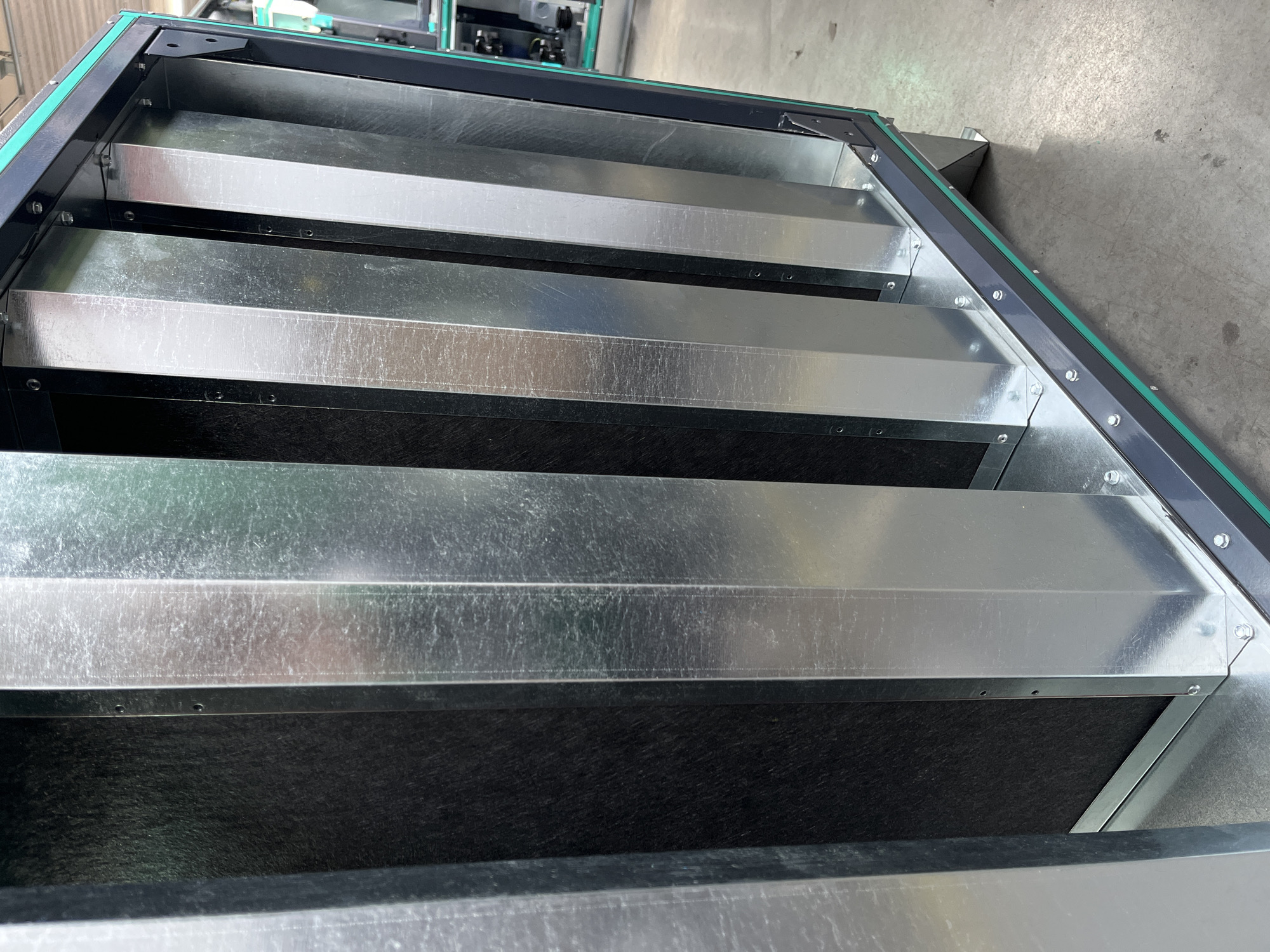 The image shows a close-up view of industrial air filtration equipment, specifically a multi-stage filter unit with several parallel, U-shaped galvanized metal channels set against a dark, fibrous filter material. The unit is housed within a dark grey metal frame with green trim. The background appears to be an indoor industrial setting with a concrete floor. There are no people or discernible landmarks that would indicate a specific location in Kyiv, Ukraine. The focus is entirely on the machinery.