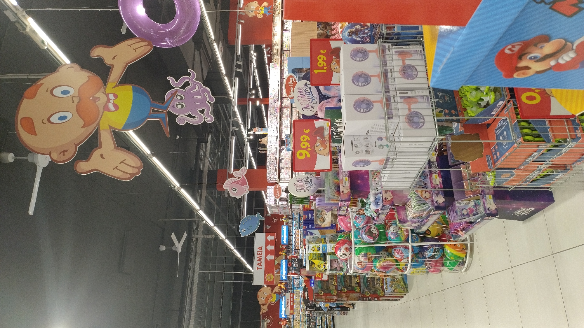 The image depicts the interior of a toy store, likely a section of a larger supermarket, located in Pylaia Municipal Unit, Greece, as suggested by the presence of Greek text on signage. In the foreground, shelves are filled with various toys, including stuffed animals, board games, and smaller packaged items like fans. Prominently displayed are several pricing signs, with one reading "9,99 €" and another "1,99 €". Hanging from the ceiling are decorative elements, including cartoon figures and inflatable rings, which are common in children's retail environments. The store is well-lit with overhead fluorescent lighting and ceiling fans are visible. The overall impression is a brightly lit, well-stocked retail space designed to attract shoppers, particularly families with children. There are no individuals clearly visible in the frame, but the arrangement of merchandise suggests an active retail environment.
