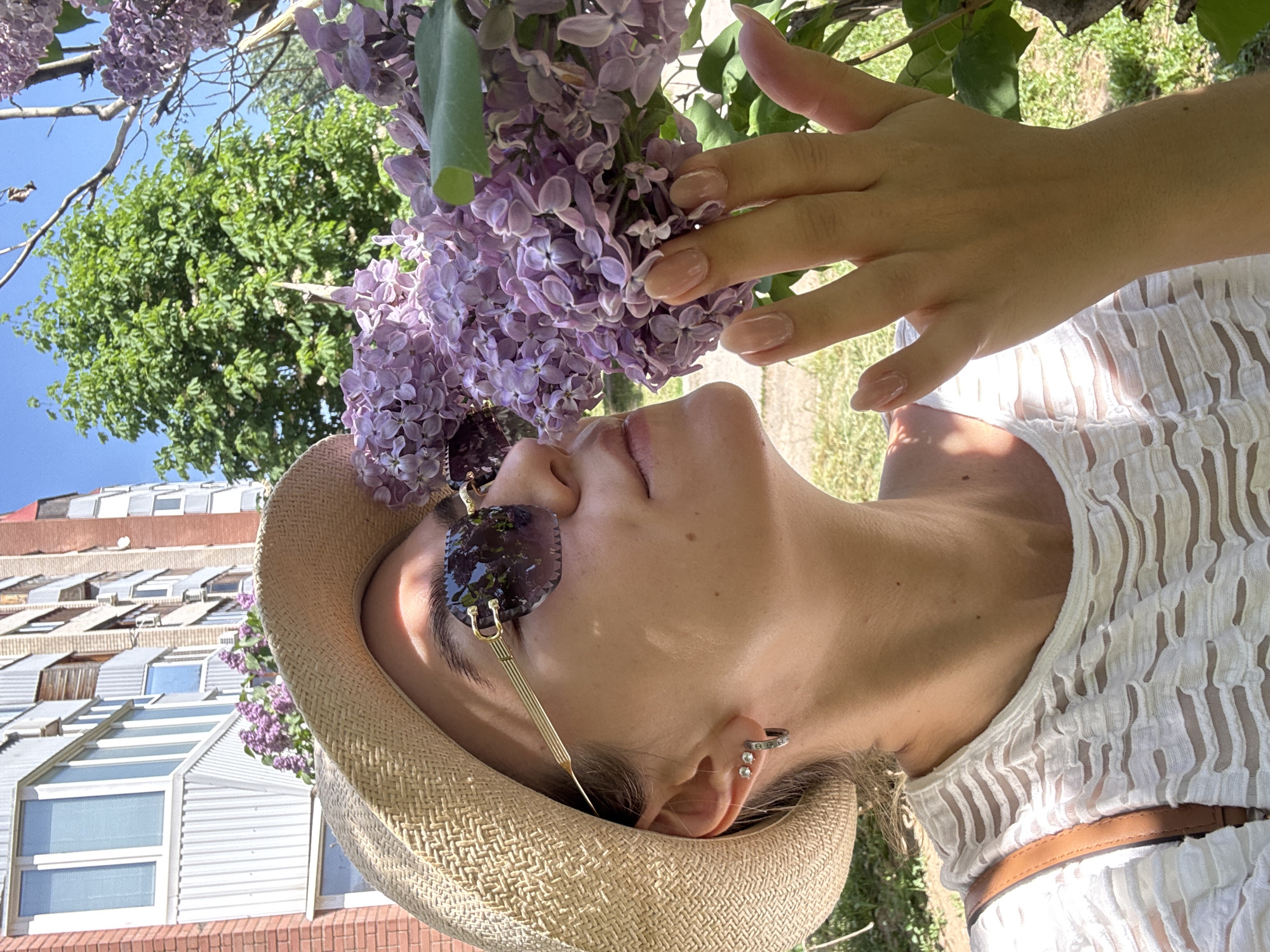 The image shows a woman, likely in her 20s or 30s, in an urban setting. She's tilted her head back and to the side, looking up and slightly to the left. She is wearing sunglasses with a gold frame and dark lenses and a straw hat. A bunch of lilacs are positioned near her face, and her hand is reaching towards them. The woman is wearing a light-colored, textured top and a brown belt. In the background, there's a building with multiple windows and an urban landscape with trees and grass. The location seems to be in the city of Unknown, Unknown.